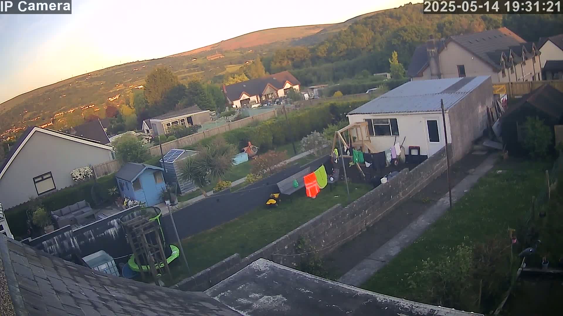 A high-angle, evening view of a residential backyard with a small shed, houses, and a hill in the background under a clear, twilight sky.