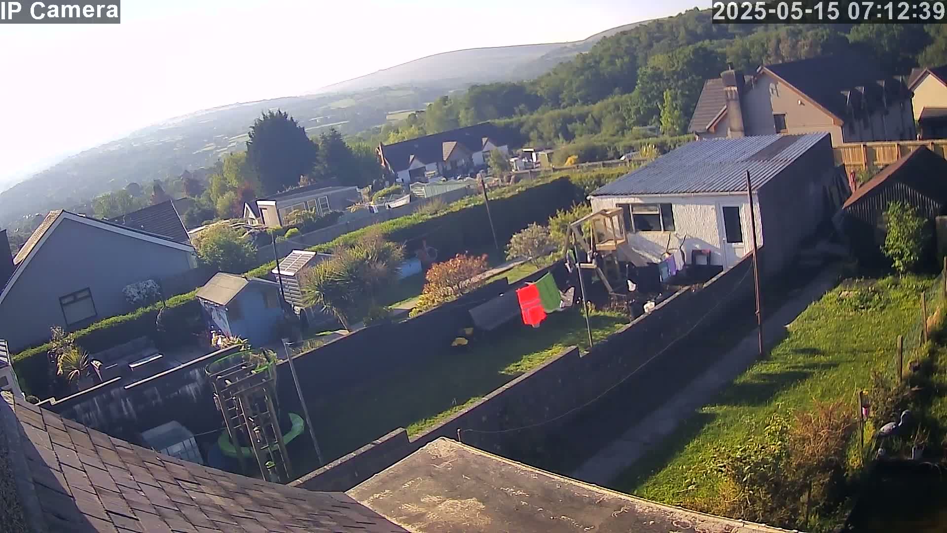A high-angle, sunny view shows several houses and gardens nestled in a hilly, green landscape.