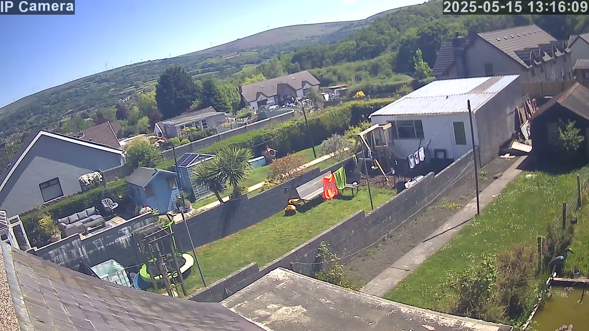 A sunny backyard with houses and a green hillside in the background shows a grassy lawn, a small pond, and several structures including a shed and a small building.
