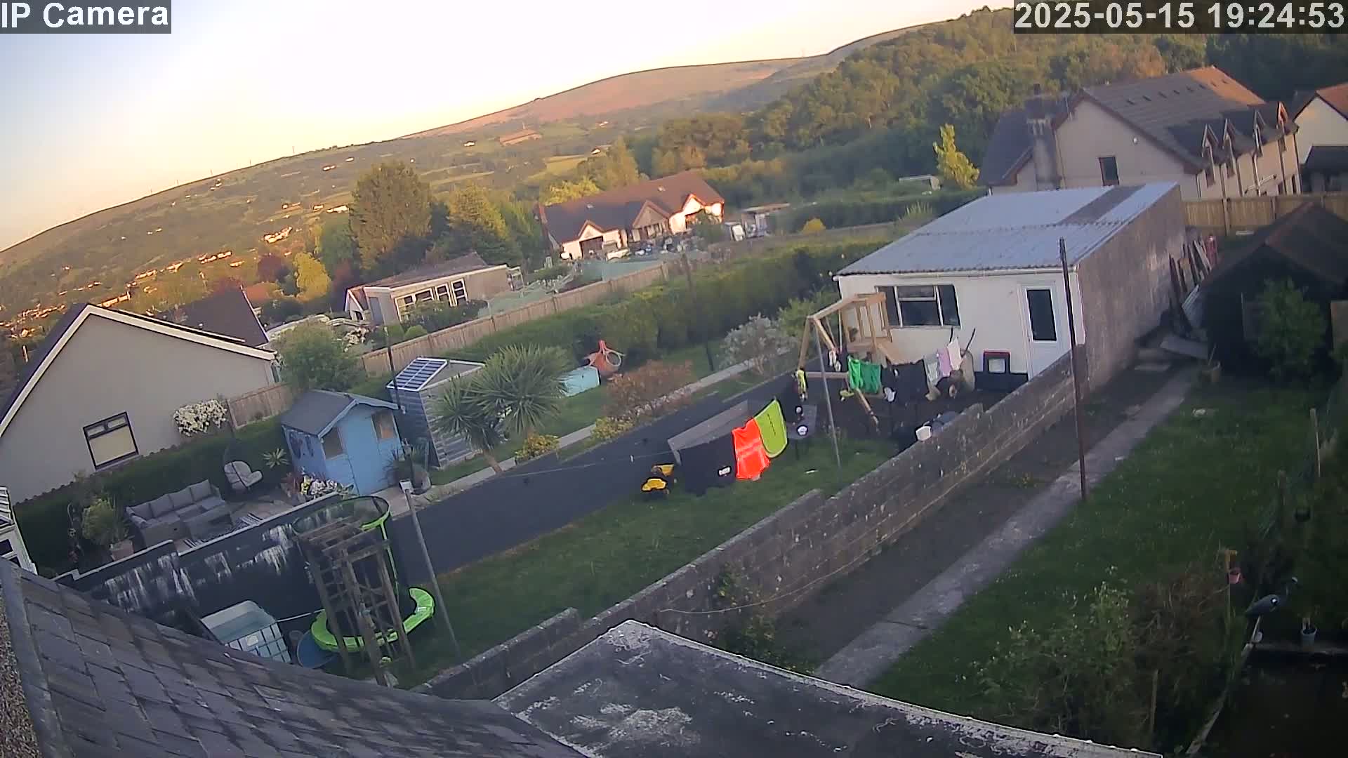 An aerial view shows residential houses and gardens in the evening, under a clear, twilight sky, with hills visible in the background.