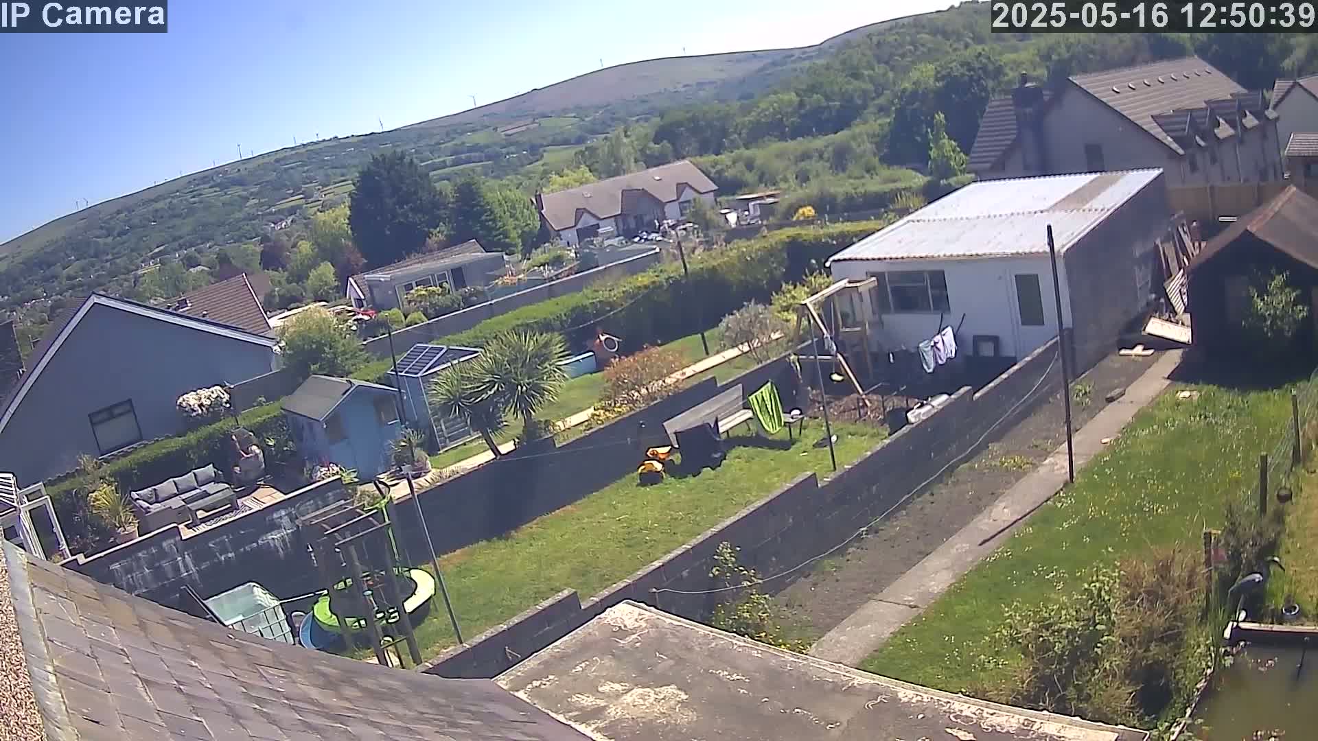 A sunny day overlooking a residential backyard with a garden, houses, and a hillside in the background.