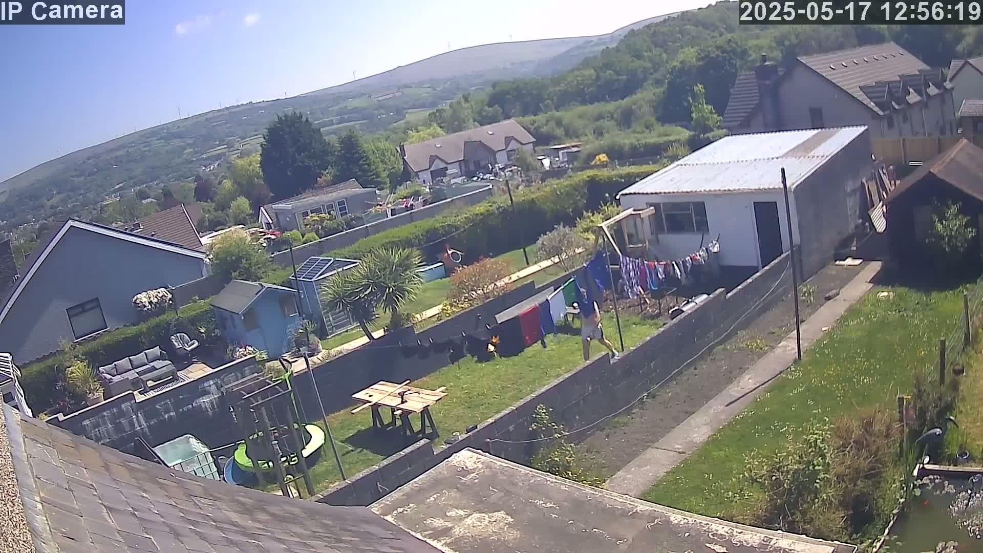 A sunny day overlooking a residential backyard with a person, a garden, several houses, and a view of distant hills.