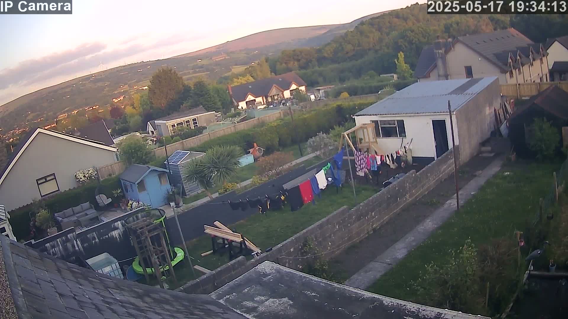 An aerial view of a residential area at dusk shows houses, gardens with clotheslines and small structures, and a hill in the background.