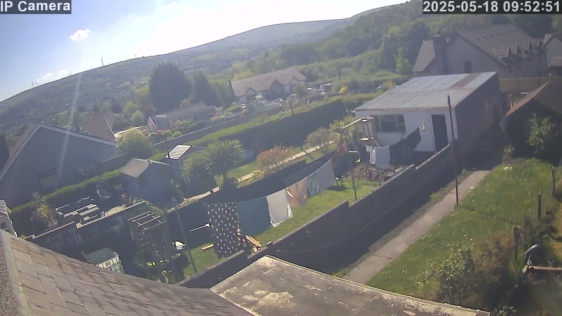 An aerial view of a residential area on a sunny day shows several houses, gardens with clotheslines, and a small shed, with wind turbines visible in the distance.