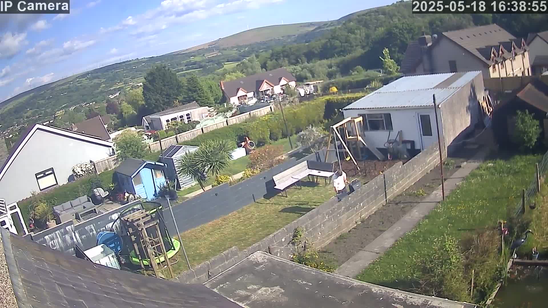 A high-angle view of a residential backyard on a partly sunny day, showing houses, gardens, and outdoor structures, including a shed, playset, and trampoline.