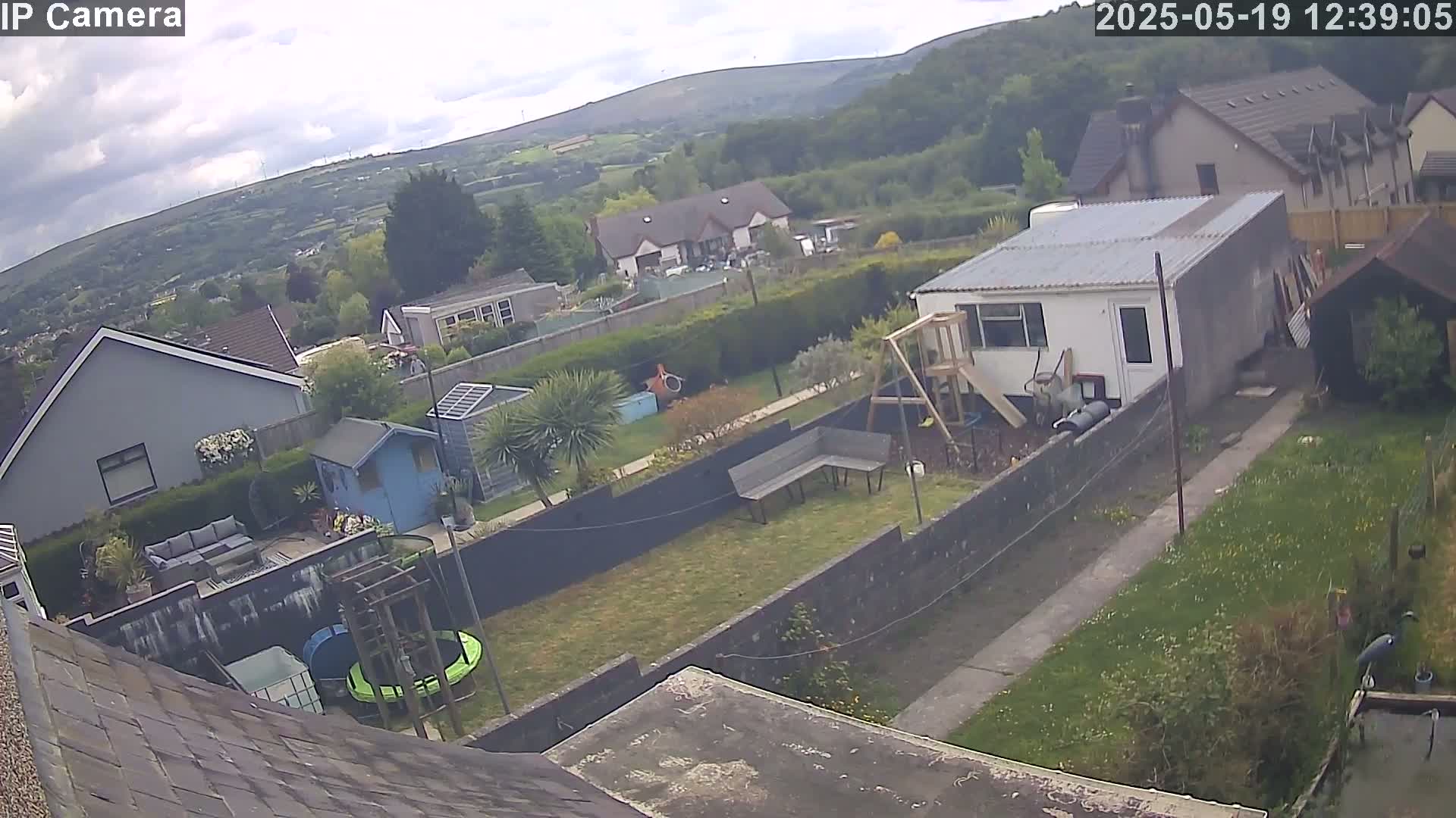 A partly cloudy day shows a backyard with a shed, a small trampoline, a playset, and a mobile home, overlooking a residential neighborhood nestled in a valley.