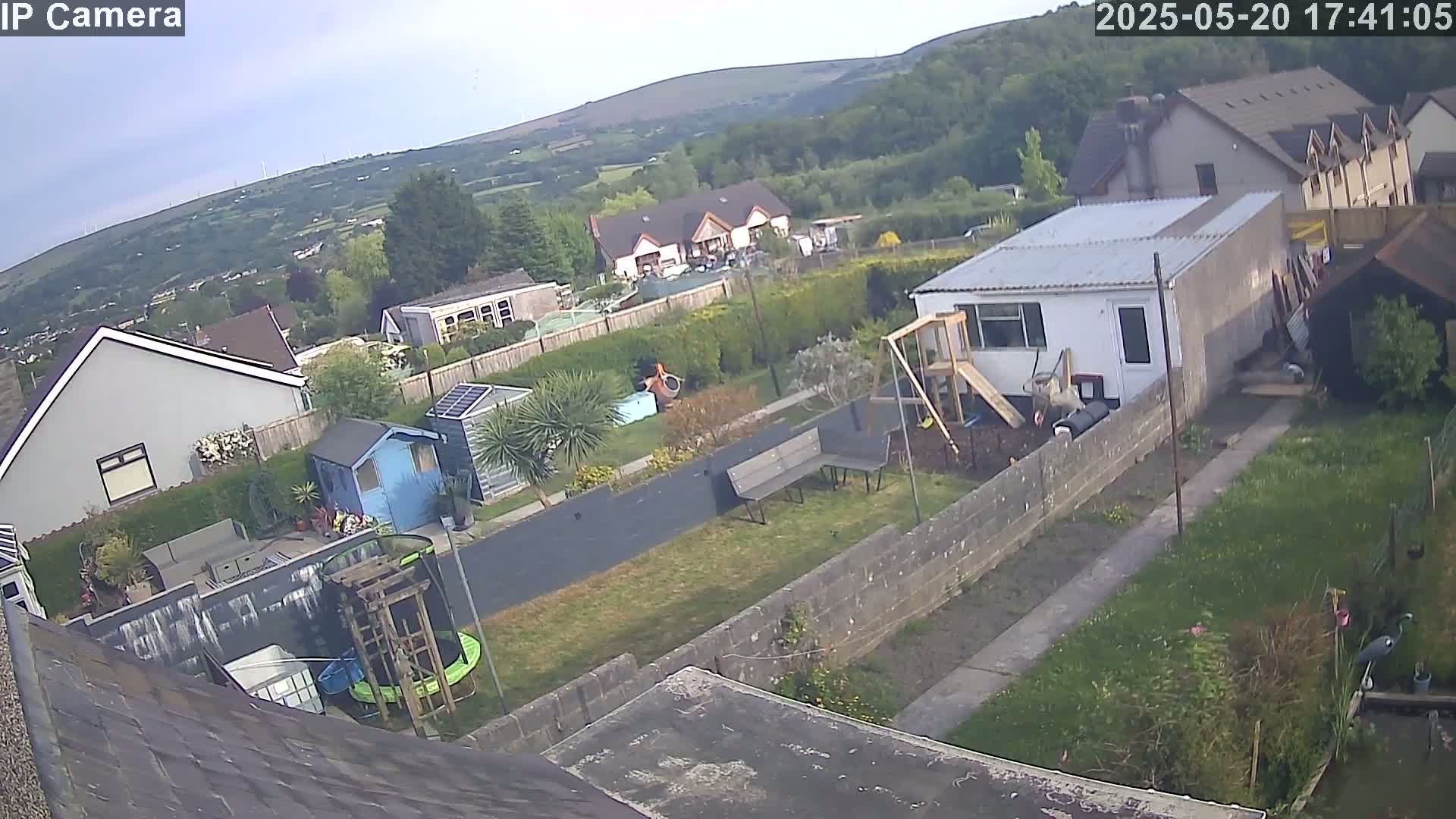 An aerial view of a backyard on a partly cloudy day shows a garden with a shed, playset, trampoline, and benches, all situated amongst houses and hills in the background.