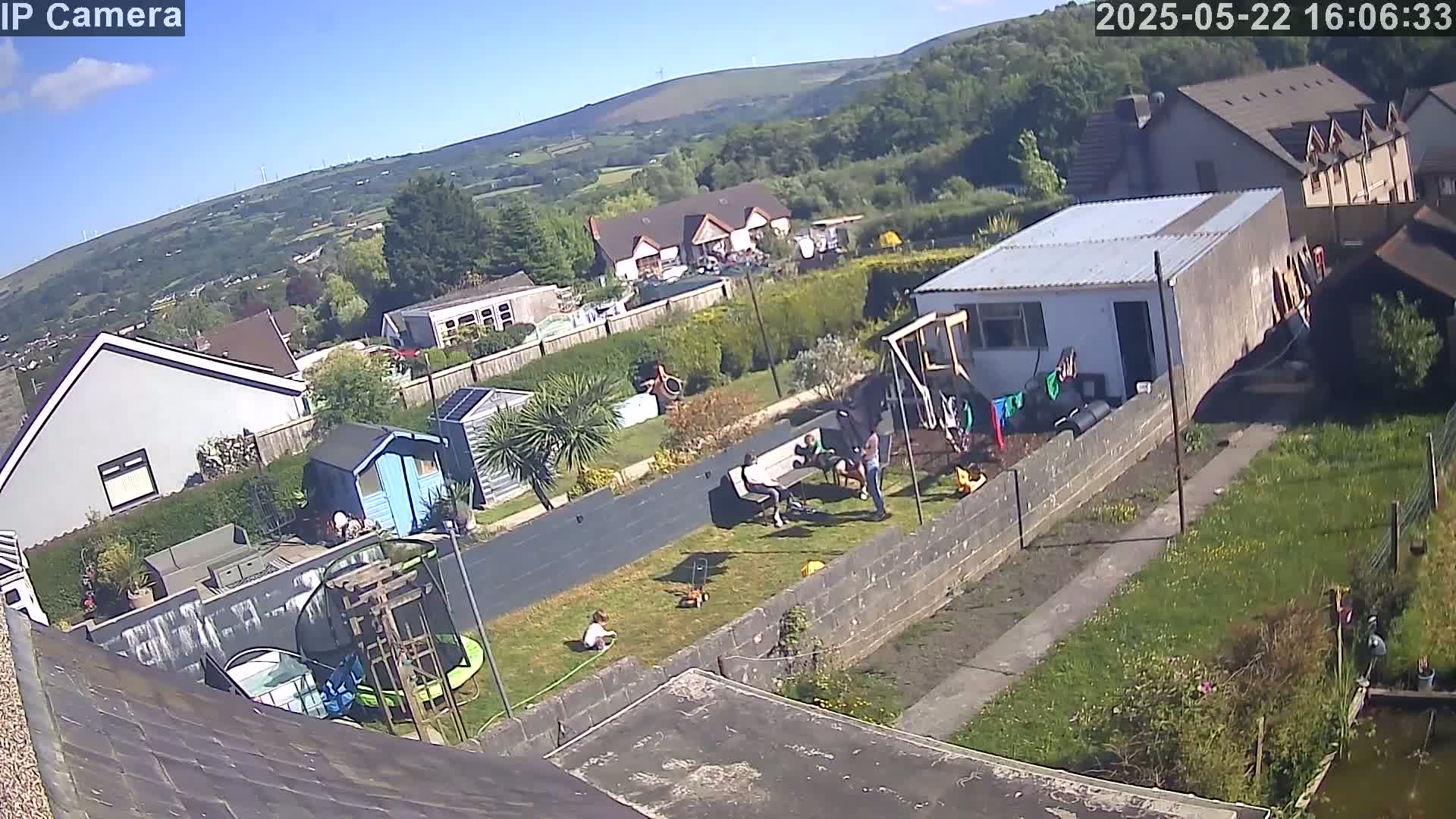 An aerial view of a backyard on a sunny day shows several people relaxing and children playing, with houses and hills visible in the background.