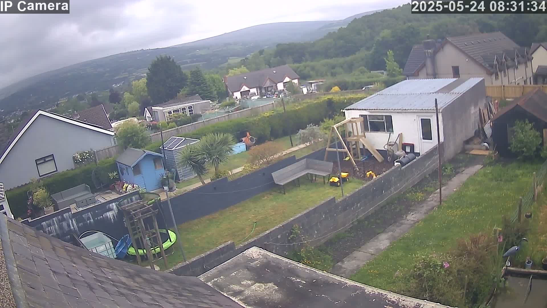 An overcast day reveals a backyard scene with a garden, several buildings including a small shed and a larger outbuilding, a playset, and a trampoline, all nestled against a backdrop of rolling hills.