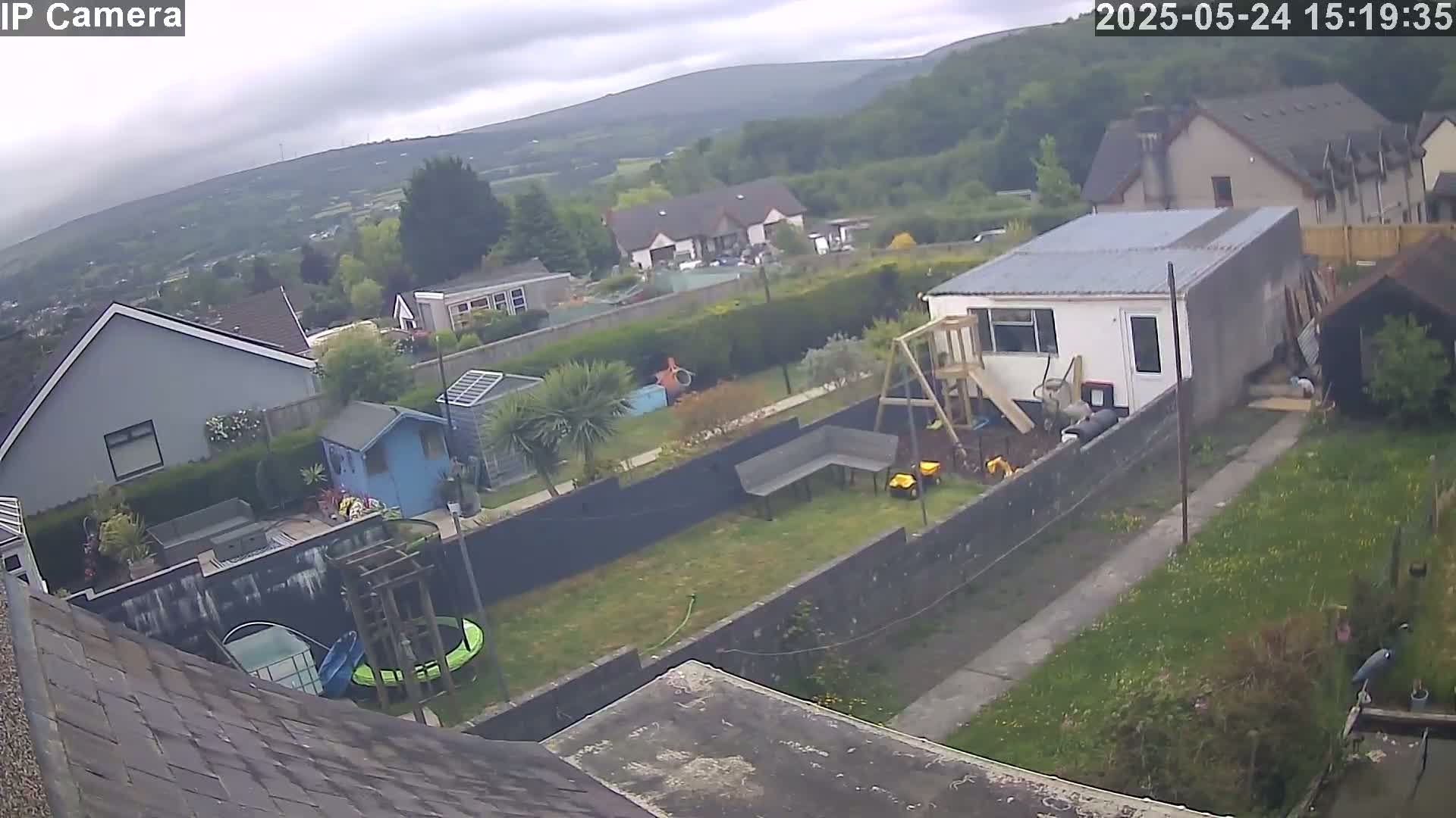 An overcast day view of a backyard with a small building, playground equipment, a garden, and houses in the background.