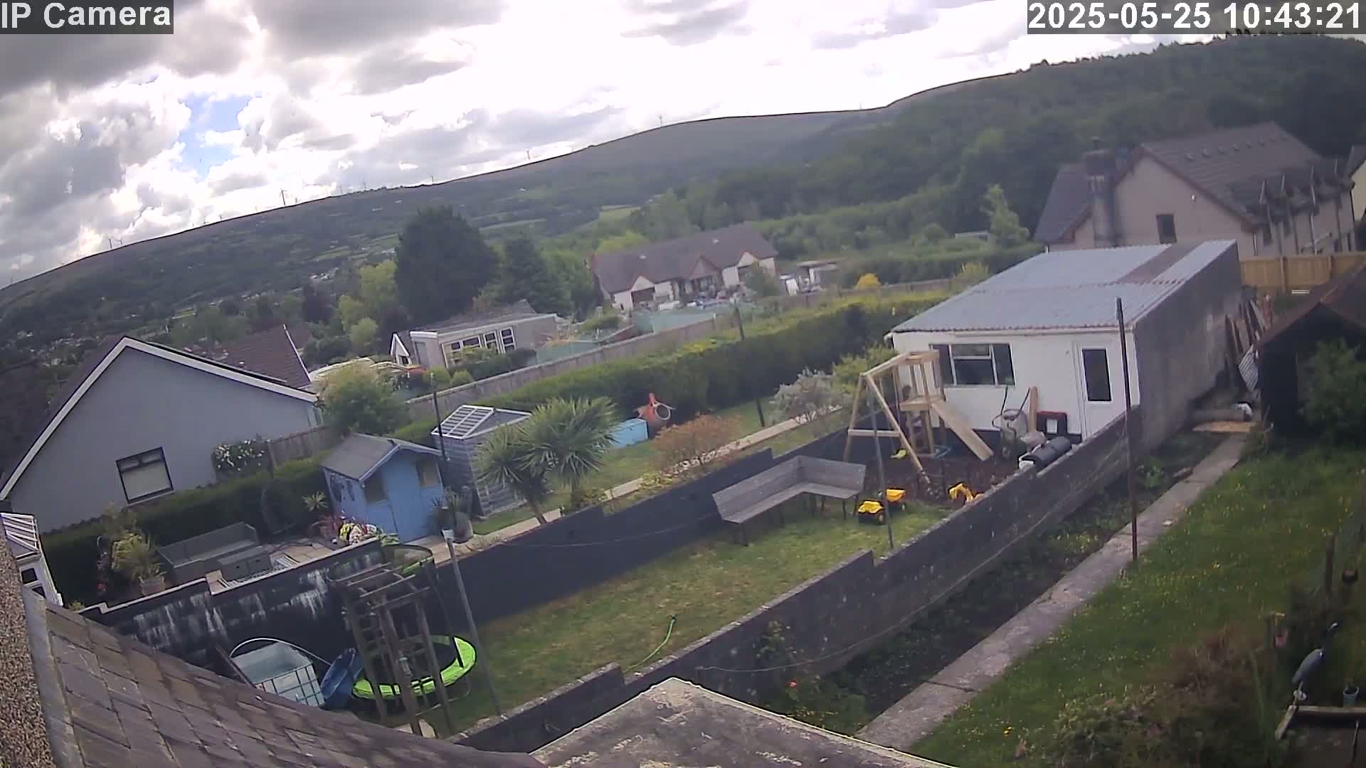 A backyard scene on a partly cloudy day shows a garden with a small shed, playset, trampoline, and a larger building, all situated amidst houses and hills in the distance.
