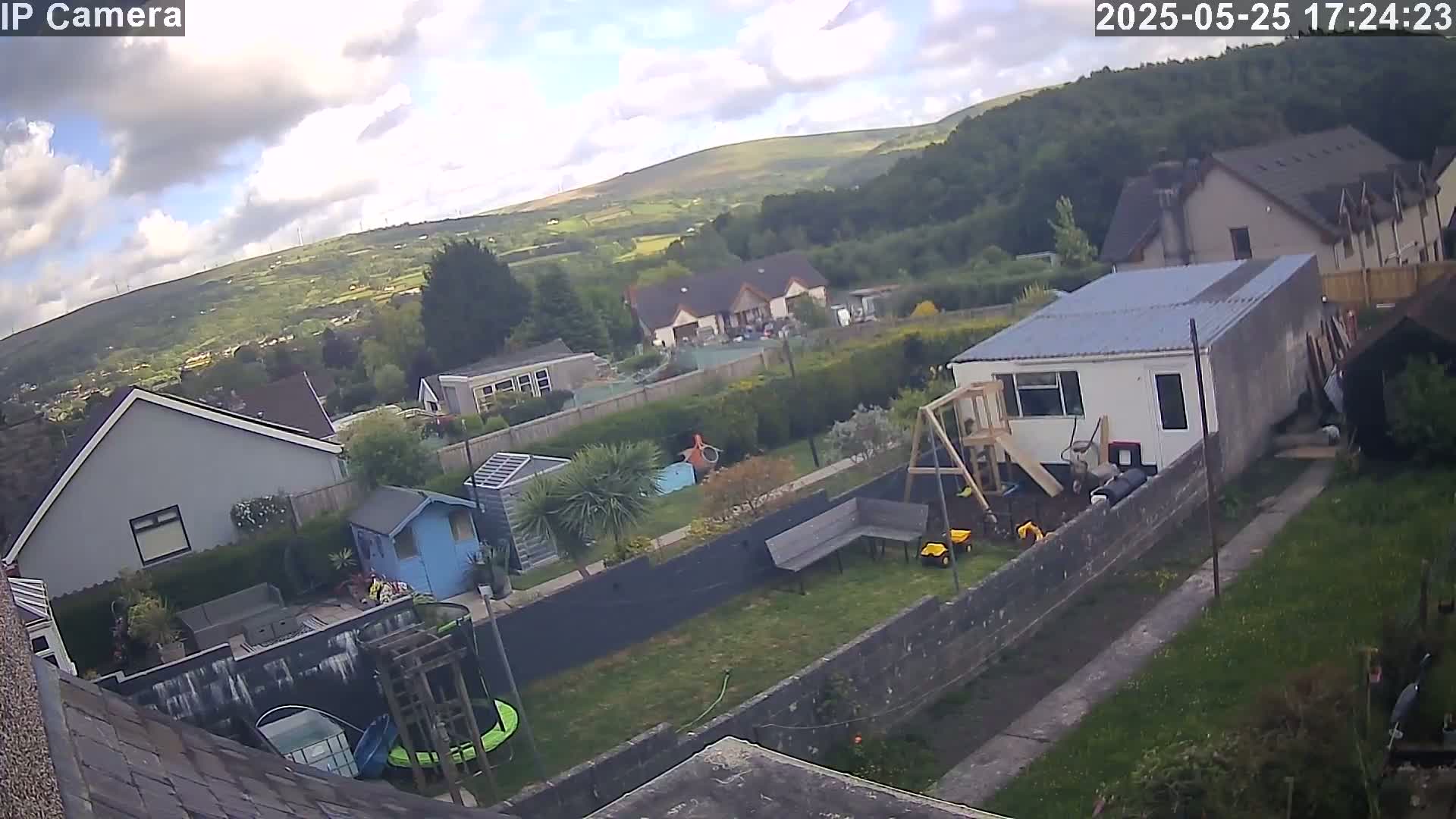 A partly cloudy afternoon view overlooking a residential backyard with a small playset and garden shed, houses, and a hill in the background.