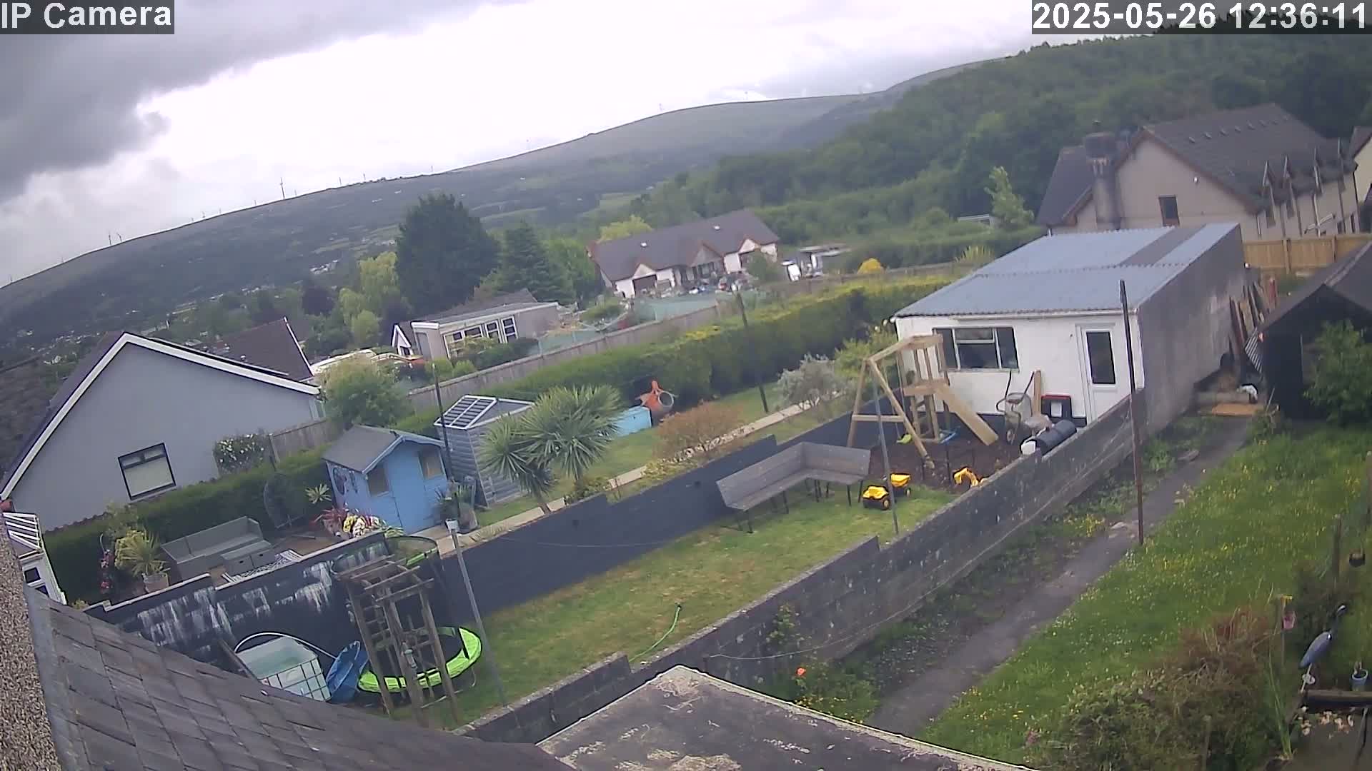 A residential backyard scene under an overcast sky shows several houses, a trampoline, a playset, and a garden, with hills and wind turbines visible in the background.