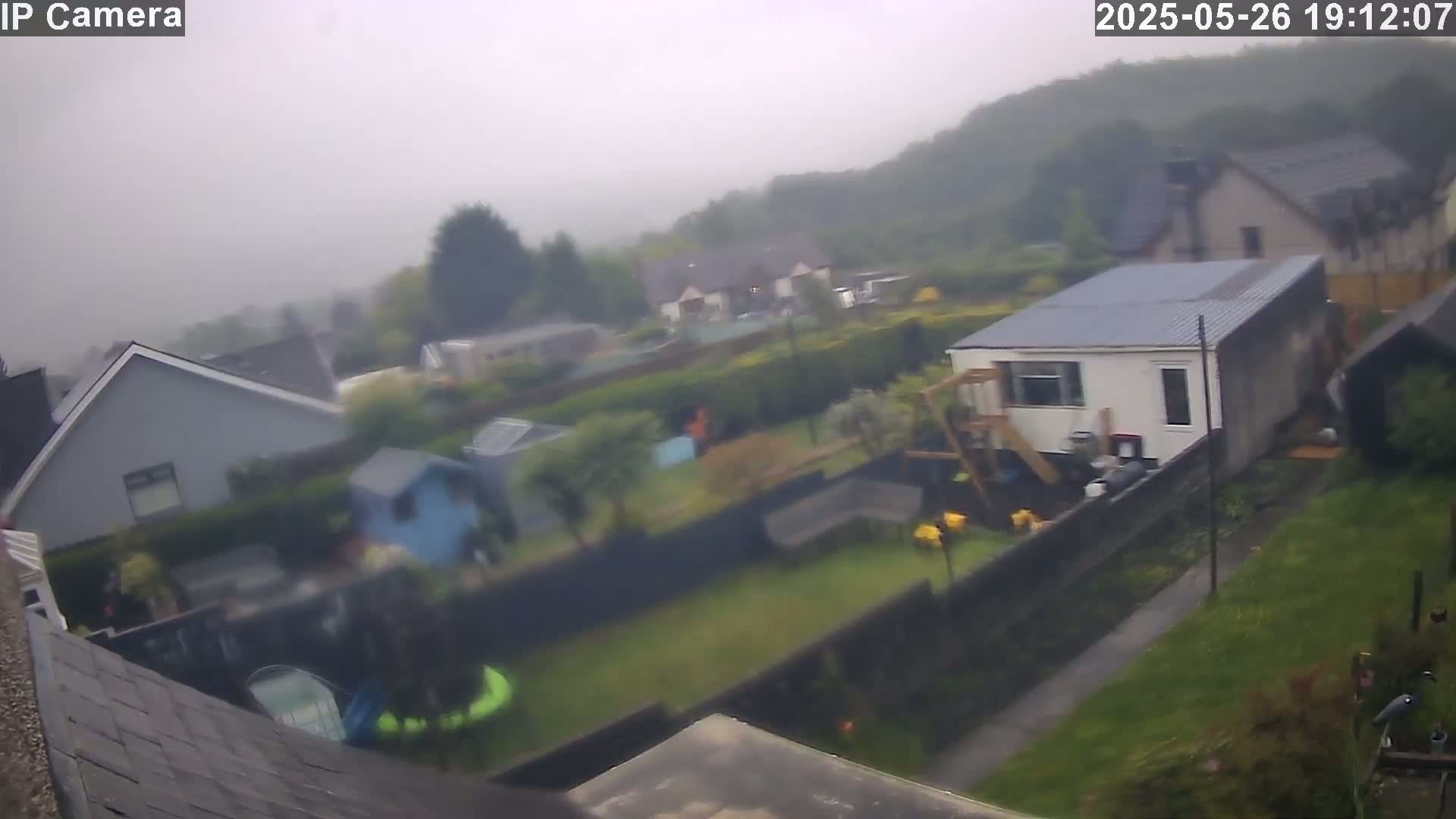 A hazy, rainy view of residential houses and gardens from a high angle, showing a backyard with a shed and play equipment.