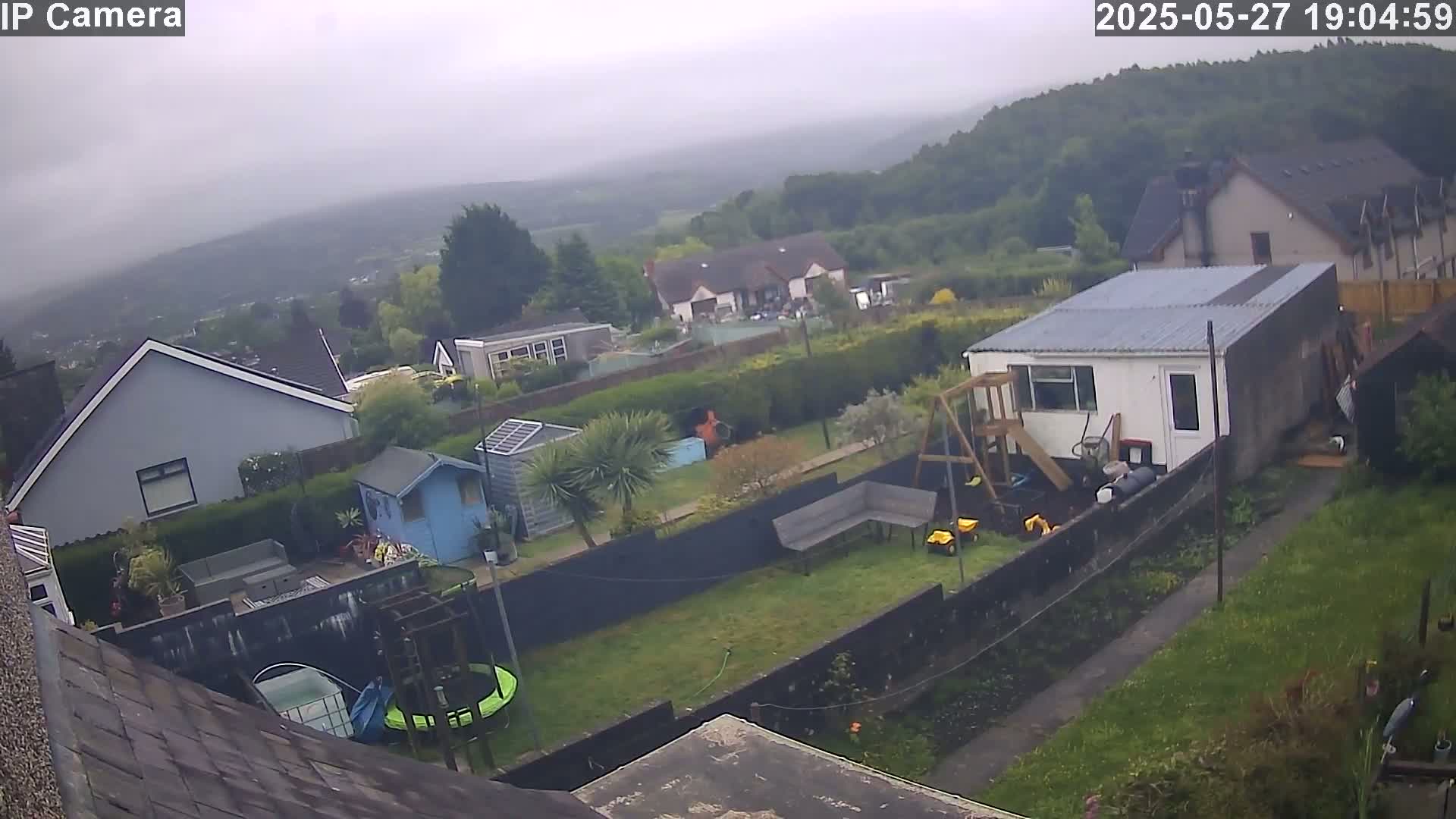 An overcast day reveals a backyard scene with houses and greenery, including a trampoline, a swing set, and various garden sheds, all situated on a hill overlooking a valley.