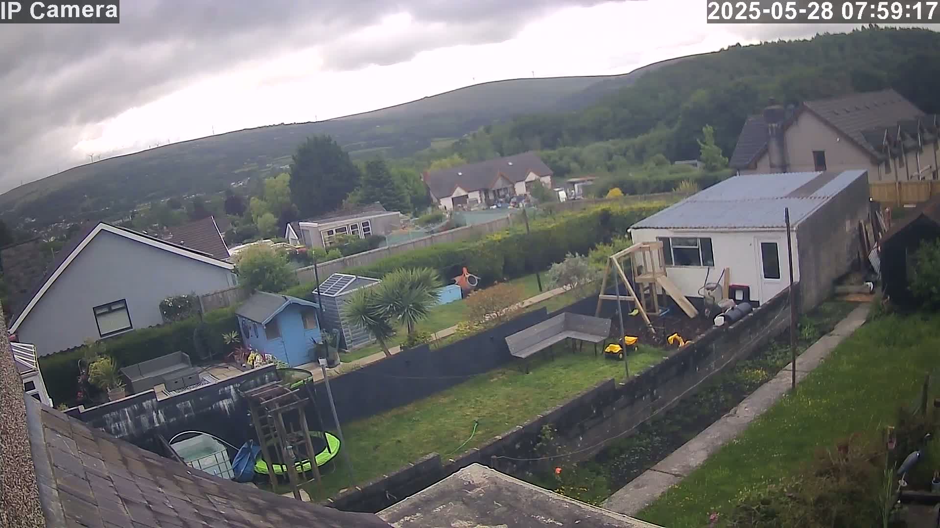 An overcast day reveals a backyard scene with houses, a shed, a playset, and a trampoline, all situated against a backdrop of green hills and a cloudy sky.