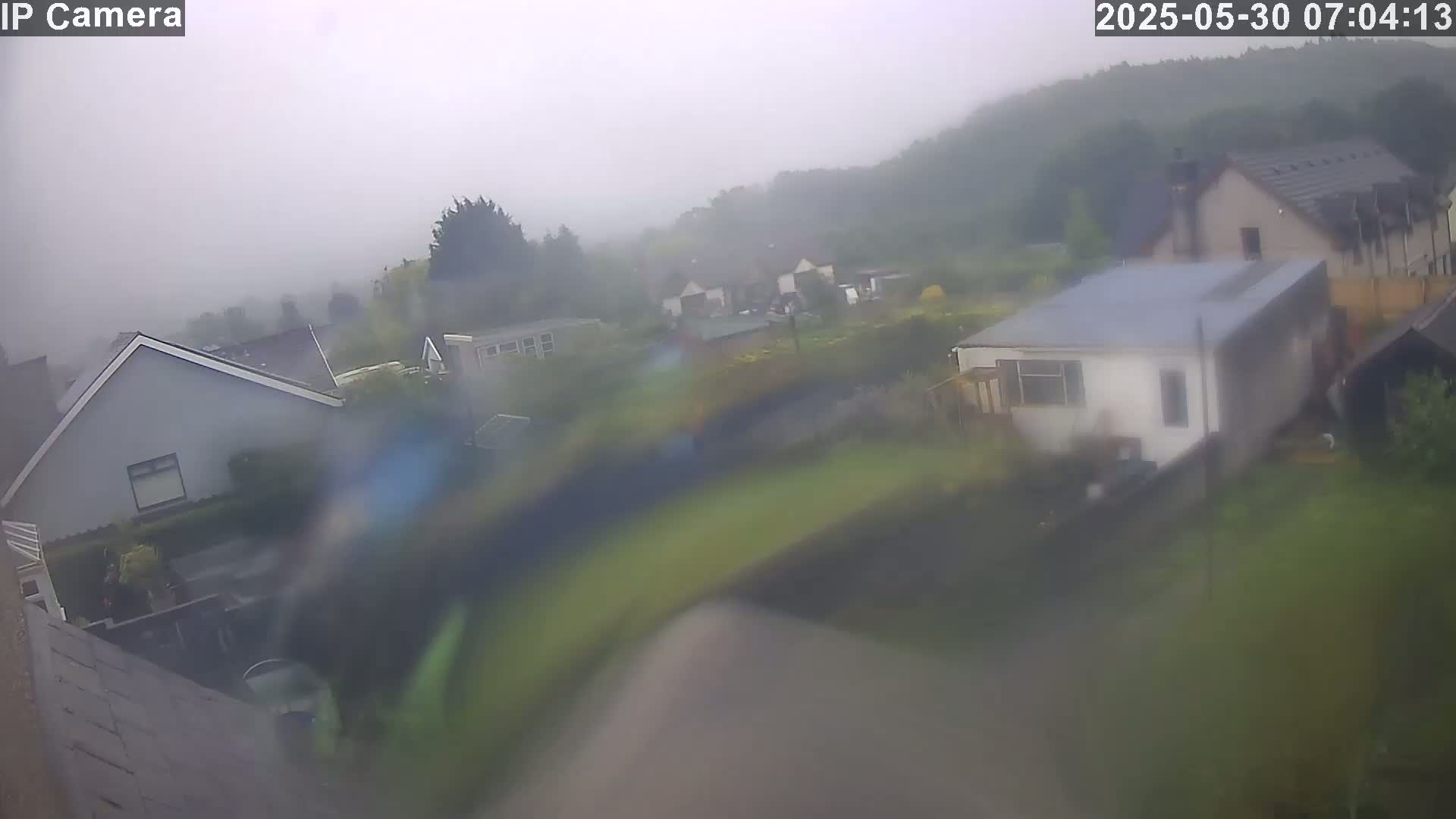 A rain-blurred view shows several houses and buildings in a residential area, with a hill covered in trees visible in the background.
