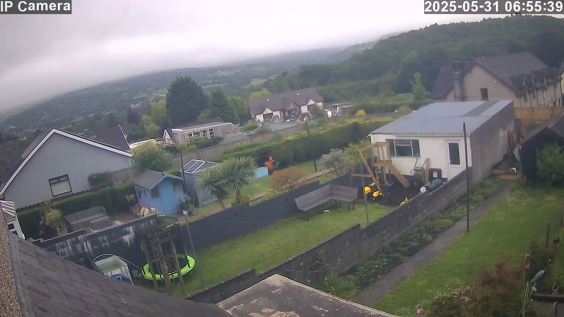 An overcast view of a residential backyard with houses and greenery, overlooking a valley in the distance.