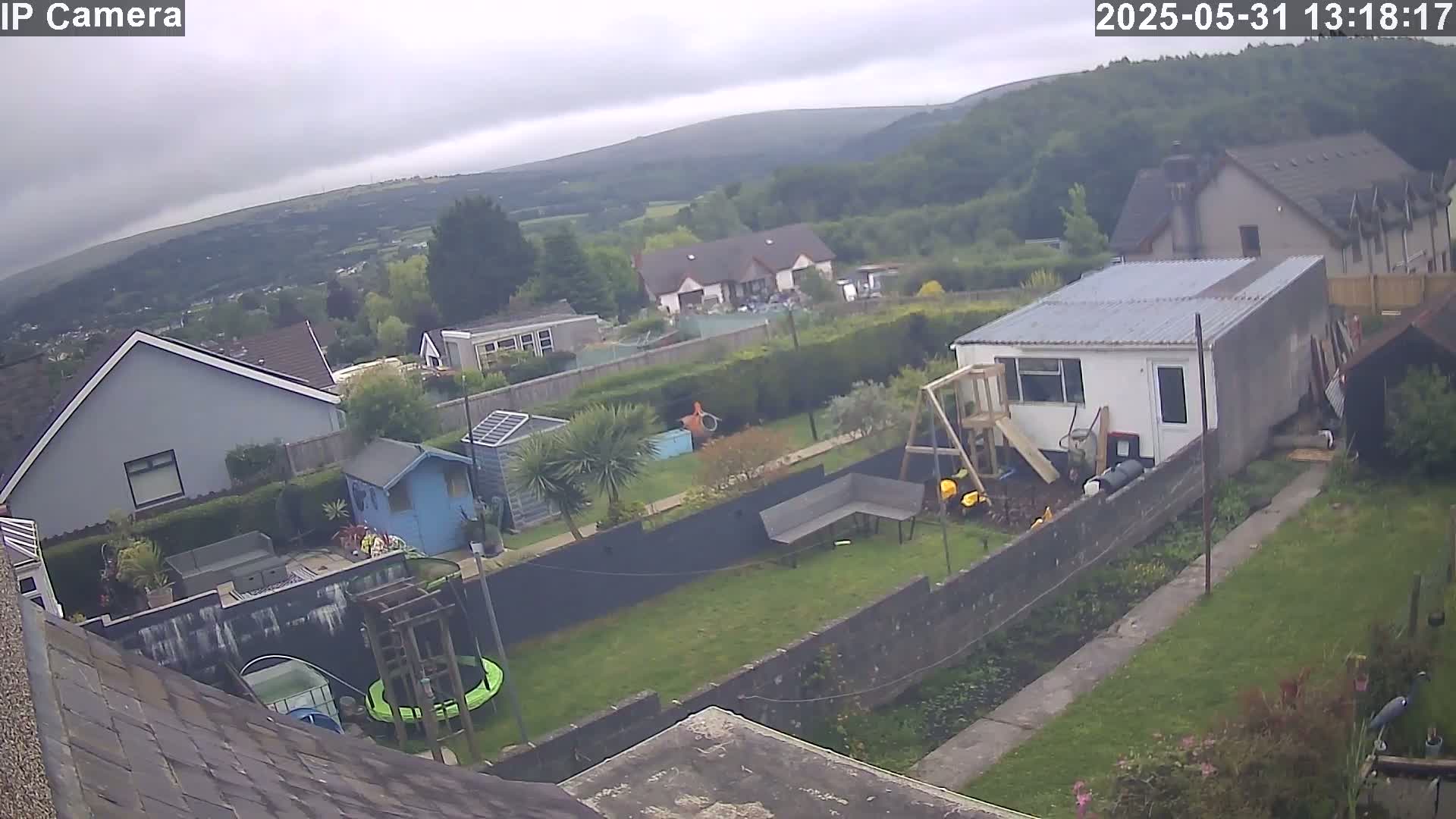 An overcast day reveals a backyard scene with houses, a garden, a trampoline, a shed, and a playset, all set against a backdrop of rolling hills.
