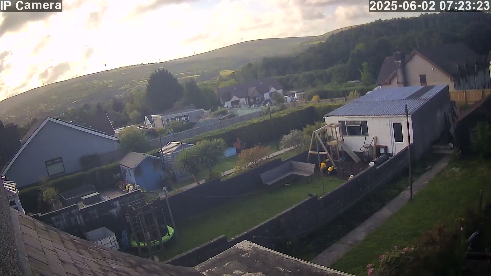 An aerial view of a backyard with houses and a hillside in the background, under partly cloudy skies.