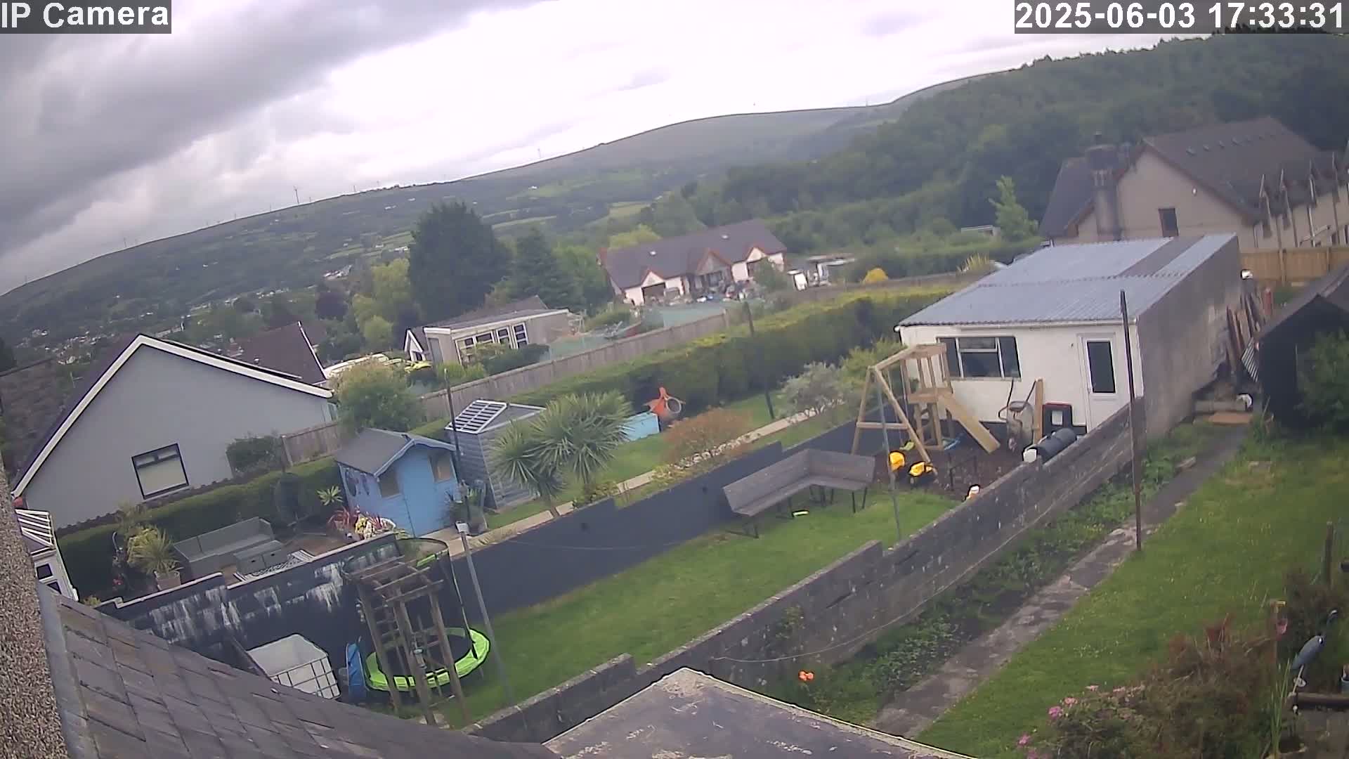 A backyard garden scene on an overcast day features houses and hills in the distance, with a trampoline, swing set, and shed visible in the foreground.