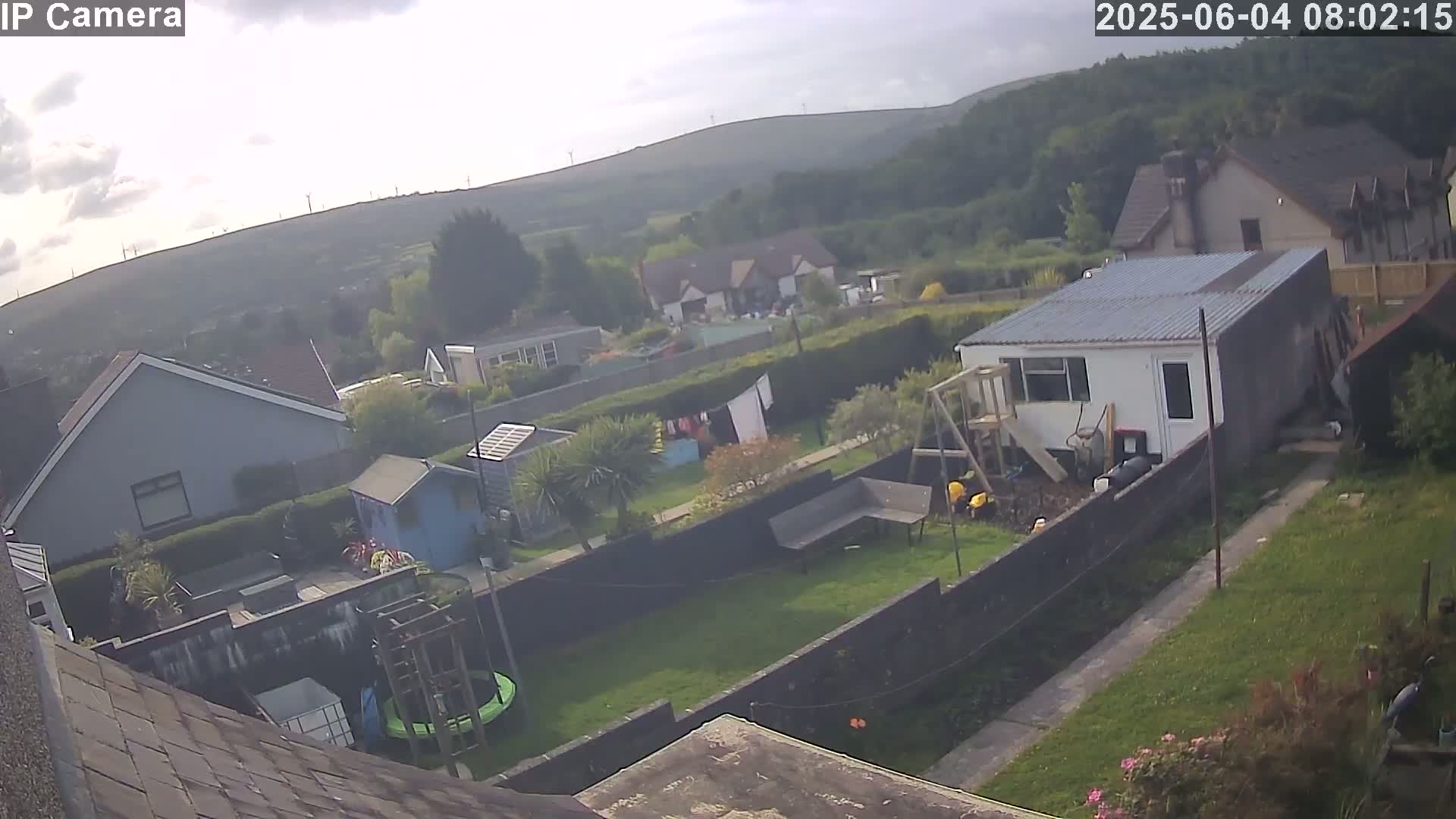 An overcast day shows a backyard with houses and hills in the background, featuring a trampoline, garden shed, and clothesline.