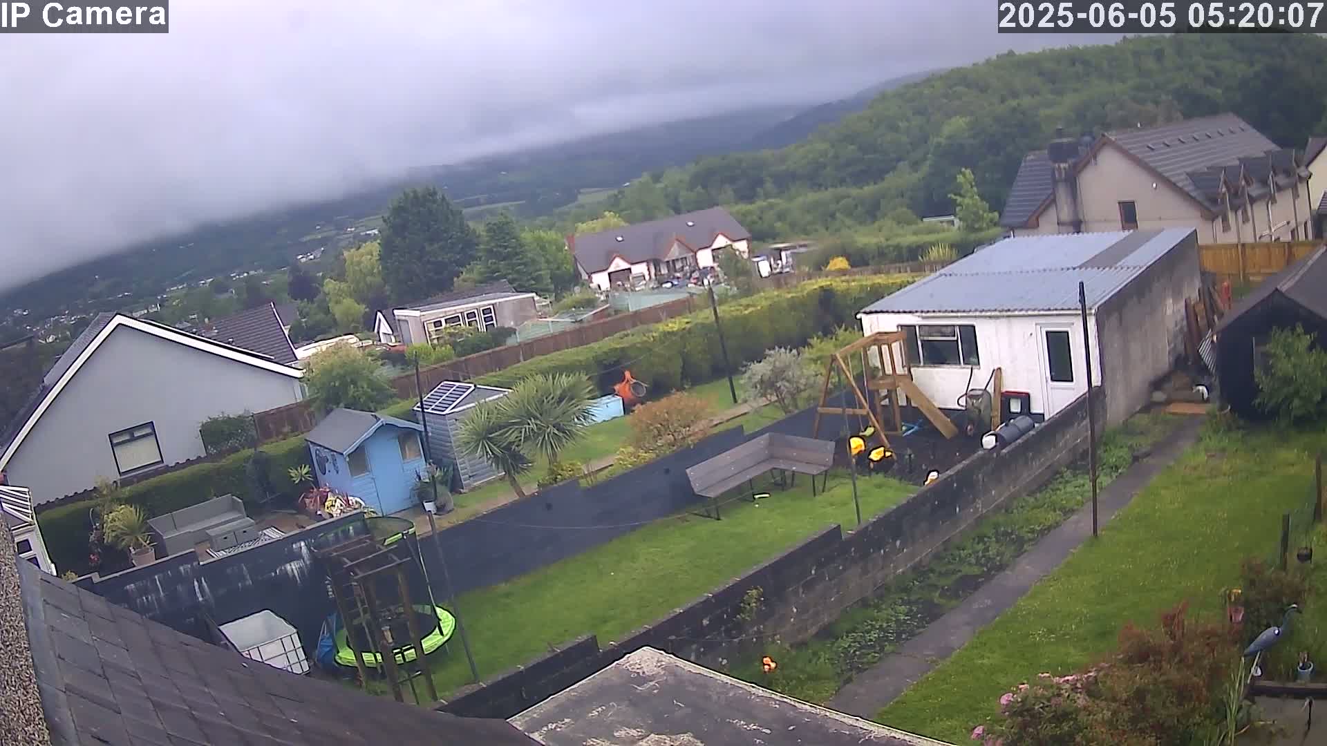 An overcast day reveals a backyard scene with houses and greenery, including a trampoline, garden shed, and playset, all nestled within a hilly landscape.