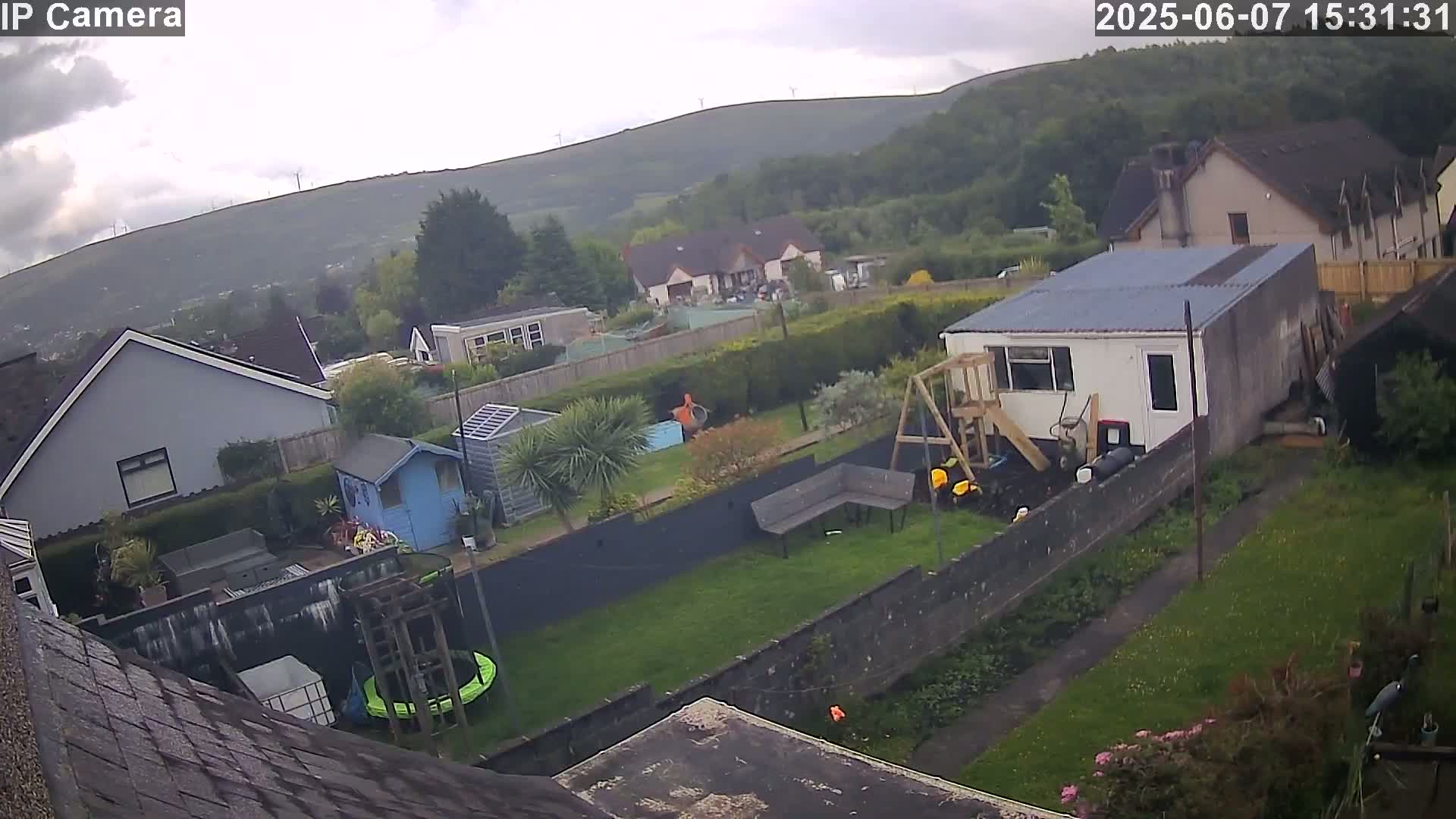 An overcast day shows a backyard with a trampoline, playset, and garden shed, bordered by houses and a hill in the distance with wind turbines visible.