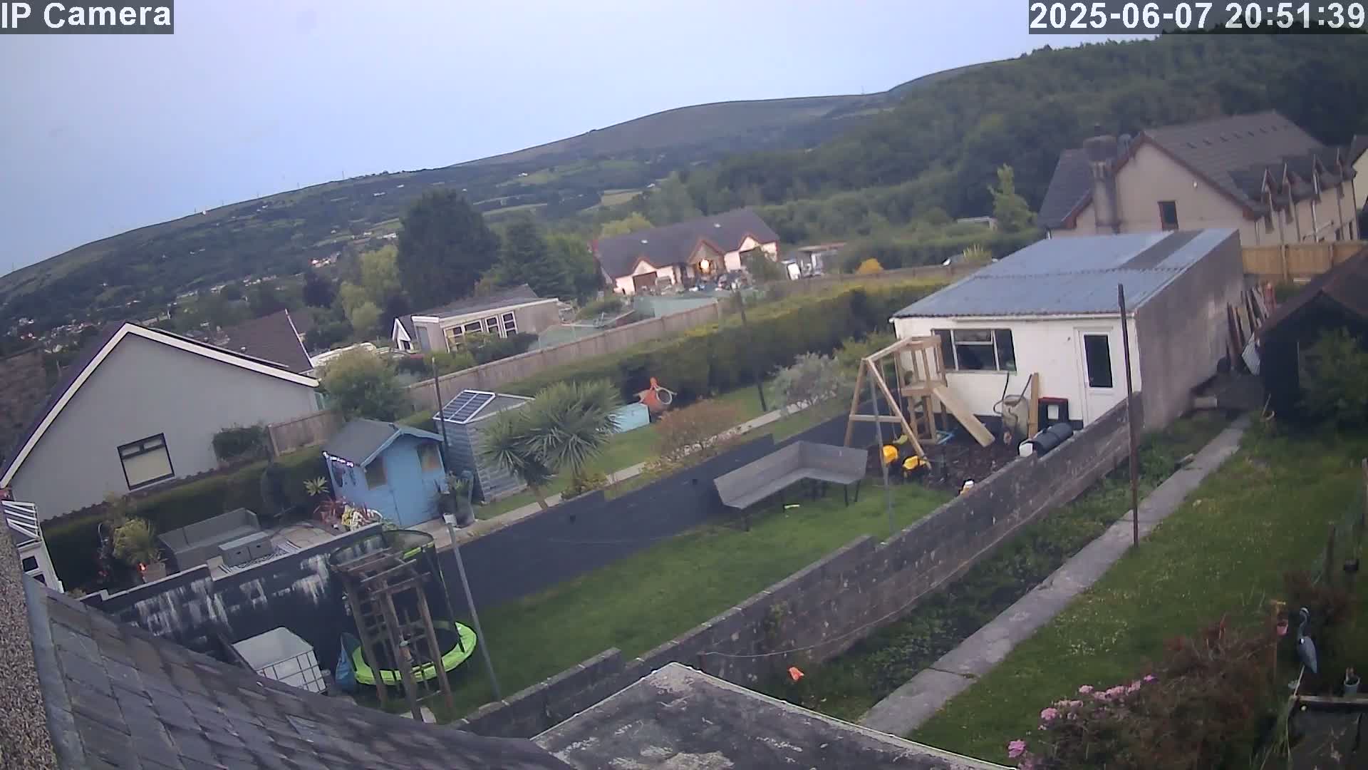 An overcast evening view of a backyard with houses and a hill in the background, featuring a garden, shed, and playset.
