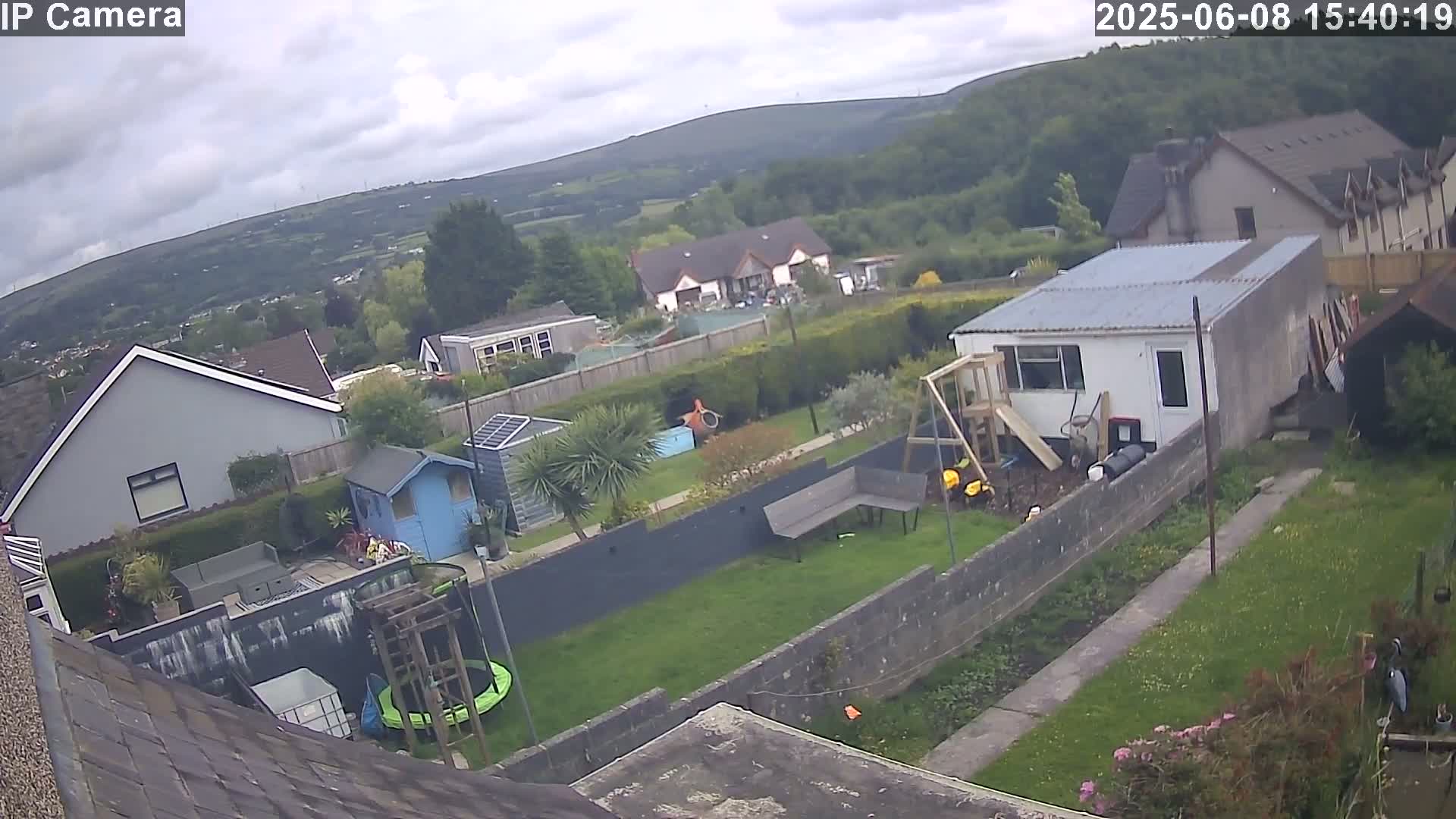 A backyard scene on a partly cloudy day shows a house, a garden with a trampoline and a small shed, and a larger building in the back, all overlooking a green valley.