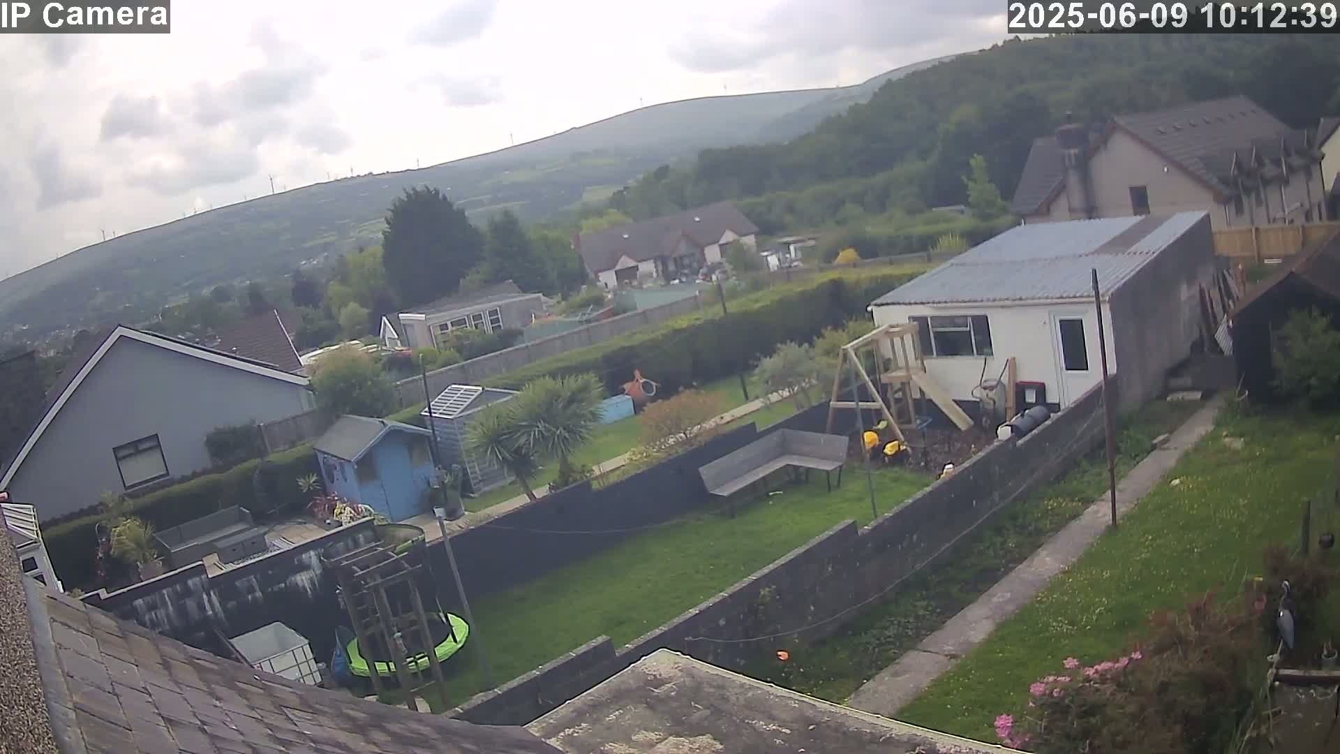 A backyard with a trampoline, garden shed, and playset overlooks houses and a hilly landscape under a cloudy sky.