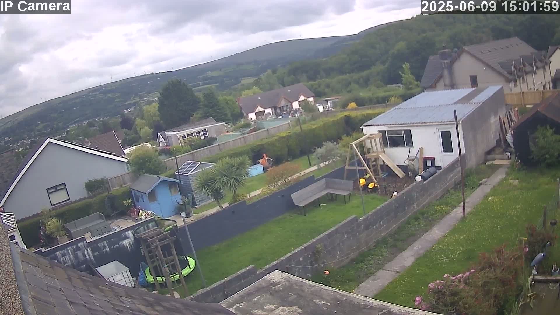An aerial view shows several houses and gardens on a cloudy day, with a small trampoline and a playset visible in one backyard.