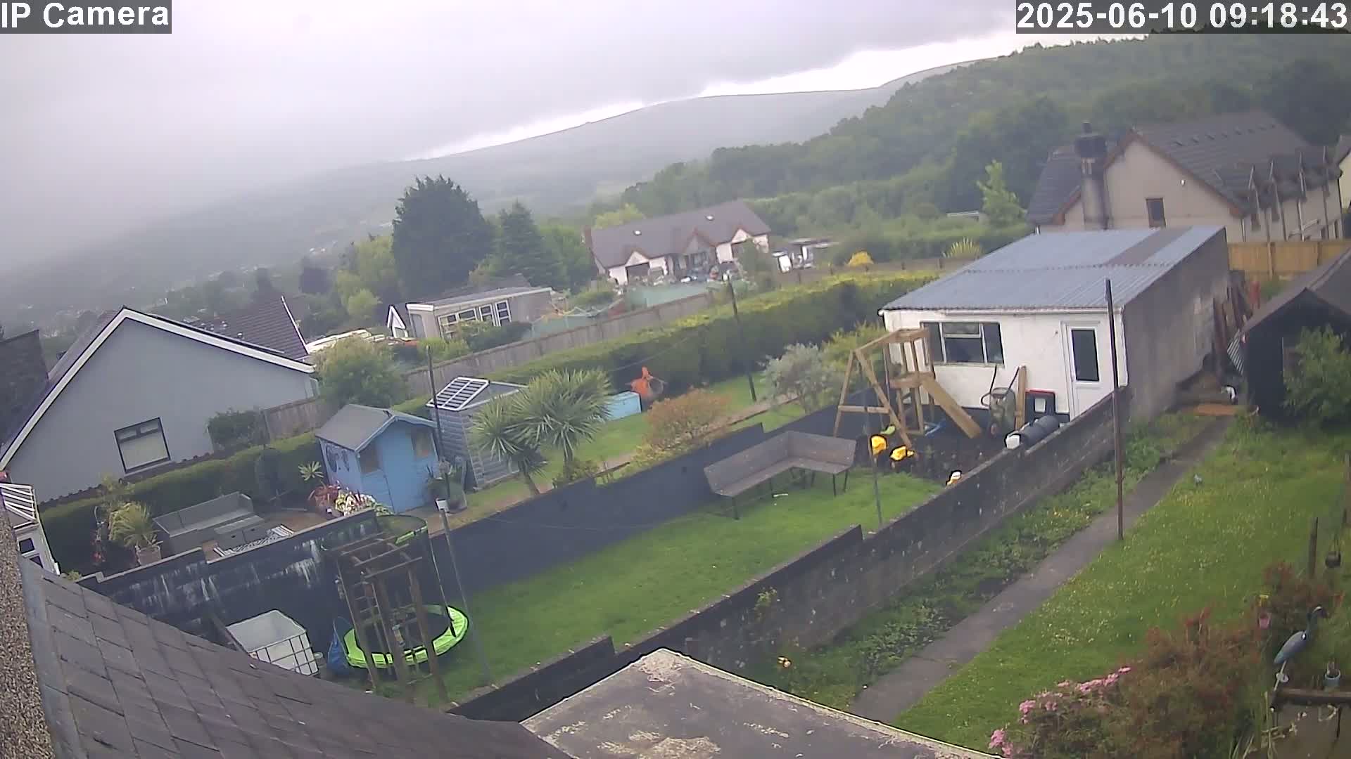 An overcast day reveals a backyard scene with houses, sheds, a trampoline, and a playset, all overlooking a hilly landscape in the distance.
