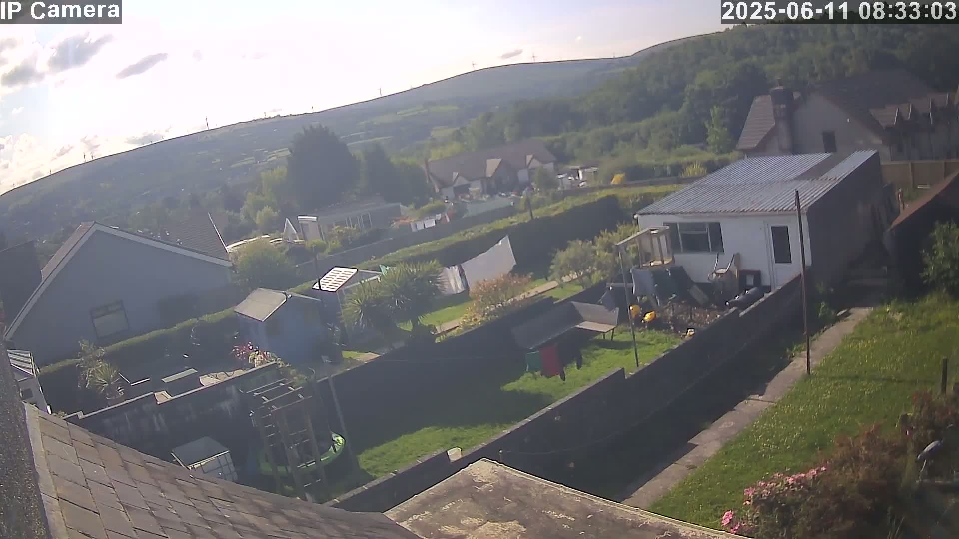 An aerial view shows a residential backyard with houses and a green hill in the background on a partly sunny day.