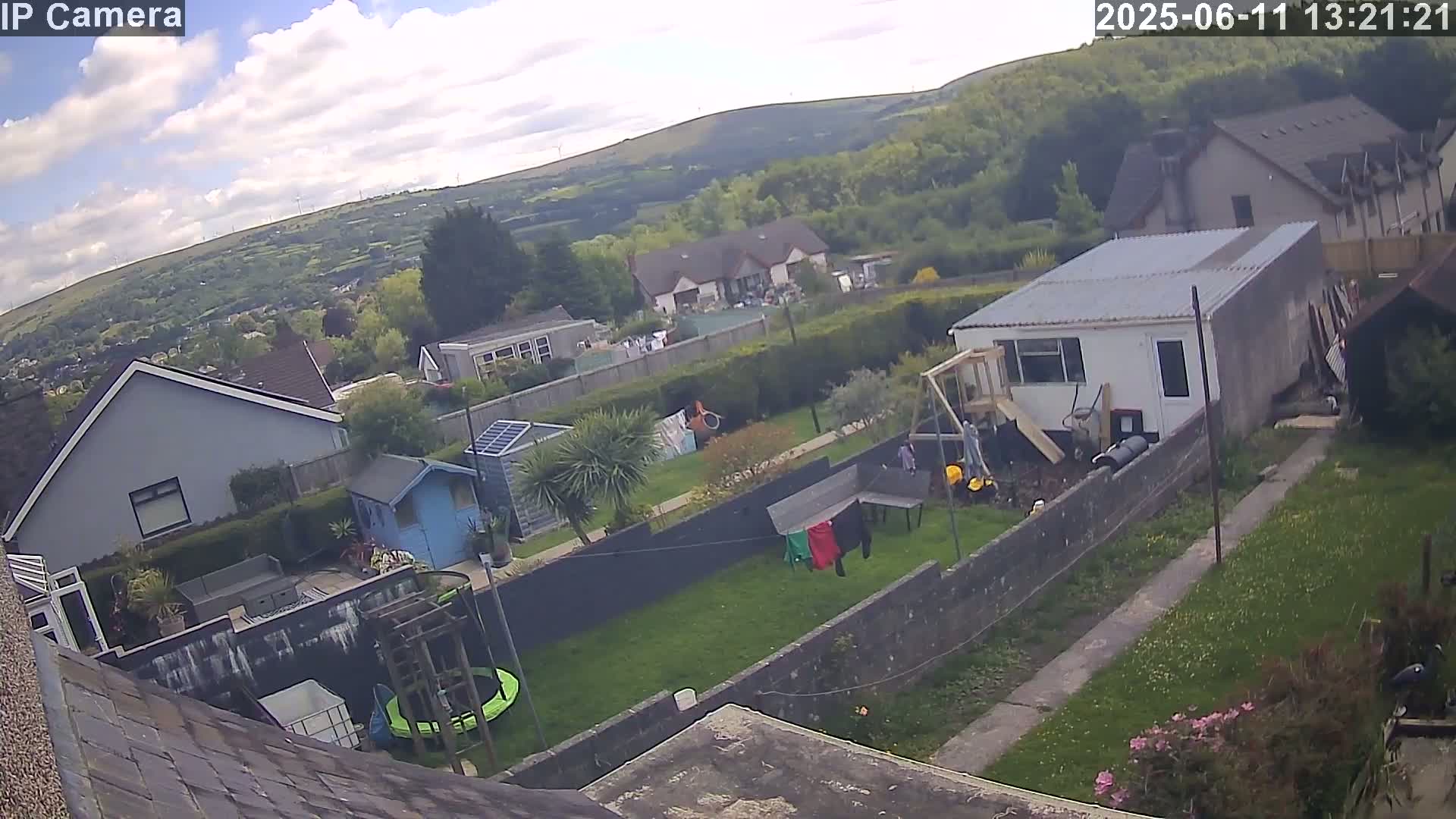 A partly cloudy day shows a view of houses and gardens, including a small trampoline and clothesline, with a hillside in the background.