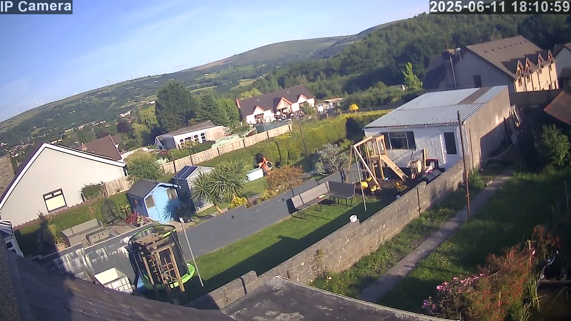 A high-angle, sunny view of residential backyards with houses, sheds, and a children's playset, overlooking a valley with wind turbines in the distance.