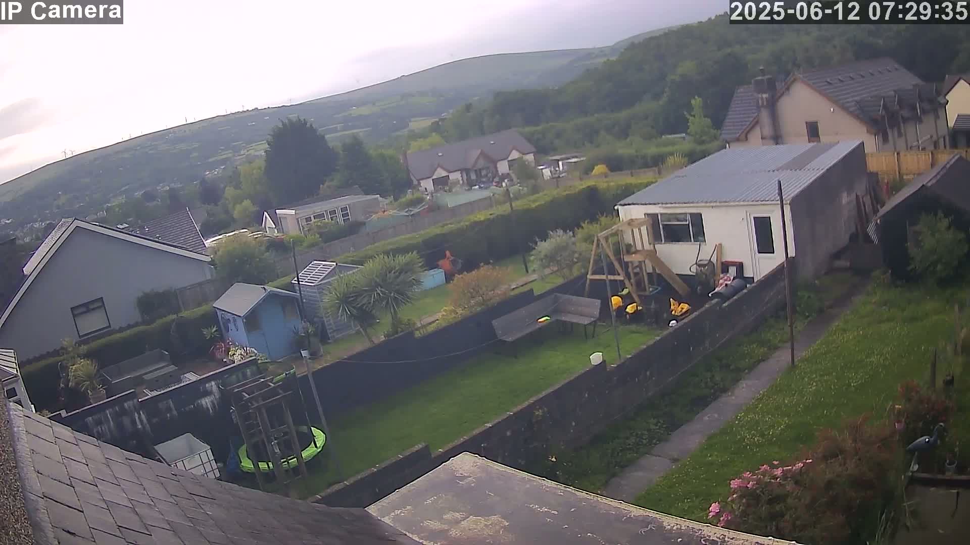 An overcast view of a backyard with a trampoline, playset, and small garden, situated in a residential area with houses and hills in the background.