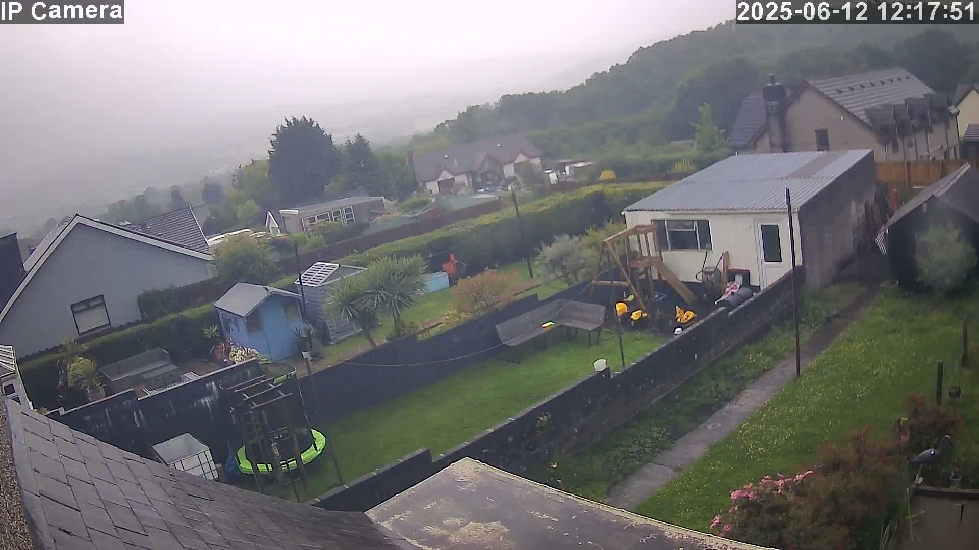 An aerial view of a residential backyard on an overcast day shows a grassy area, several outbuildings, and a trampoline, all enclosed by a fence.