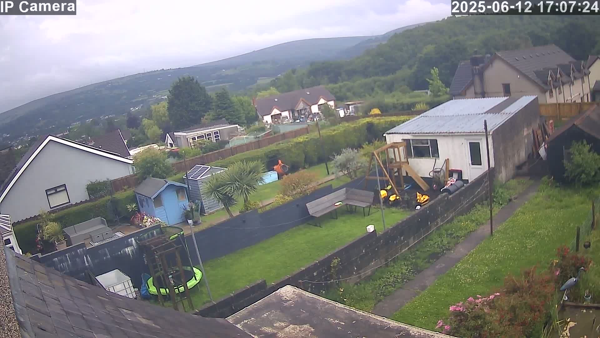 A backyard scene on an overcast day shows a garden with a trampoline, shed, playset, and a small building, all surrounded by a fence and overlooking a hilly landscape.