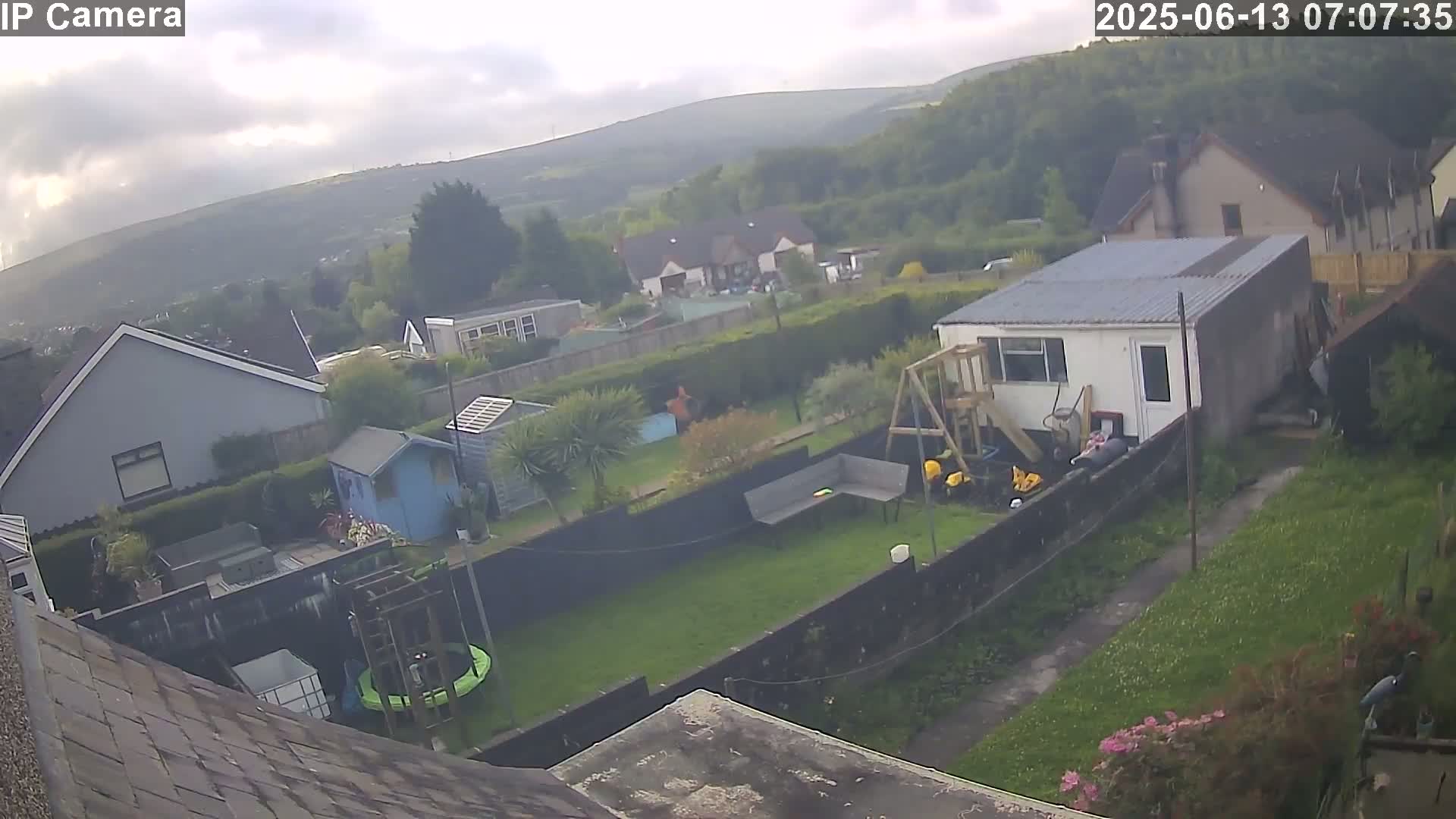 A view from a rooftop shows a residential backyard with a trampoline, garden shed, and playset, overlooking a neighborhood nestled against a hillside under a cloudy sky.
