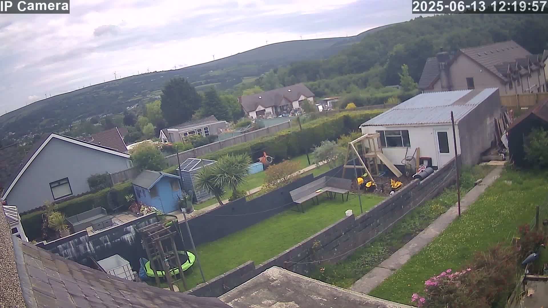An aerial view of a residential backyard on an overcast day, showing houses, a garden with a trampoline and playset, and a distant view of hills with wind turbines.