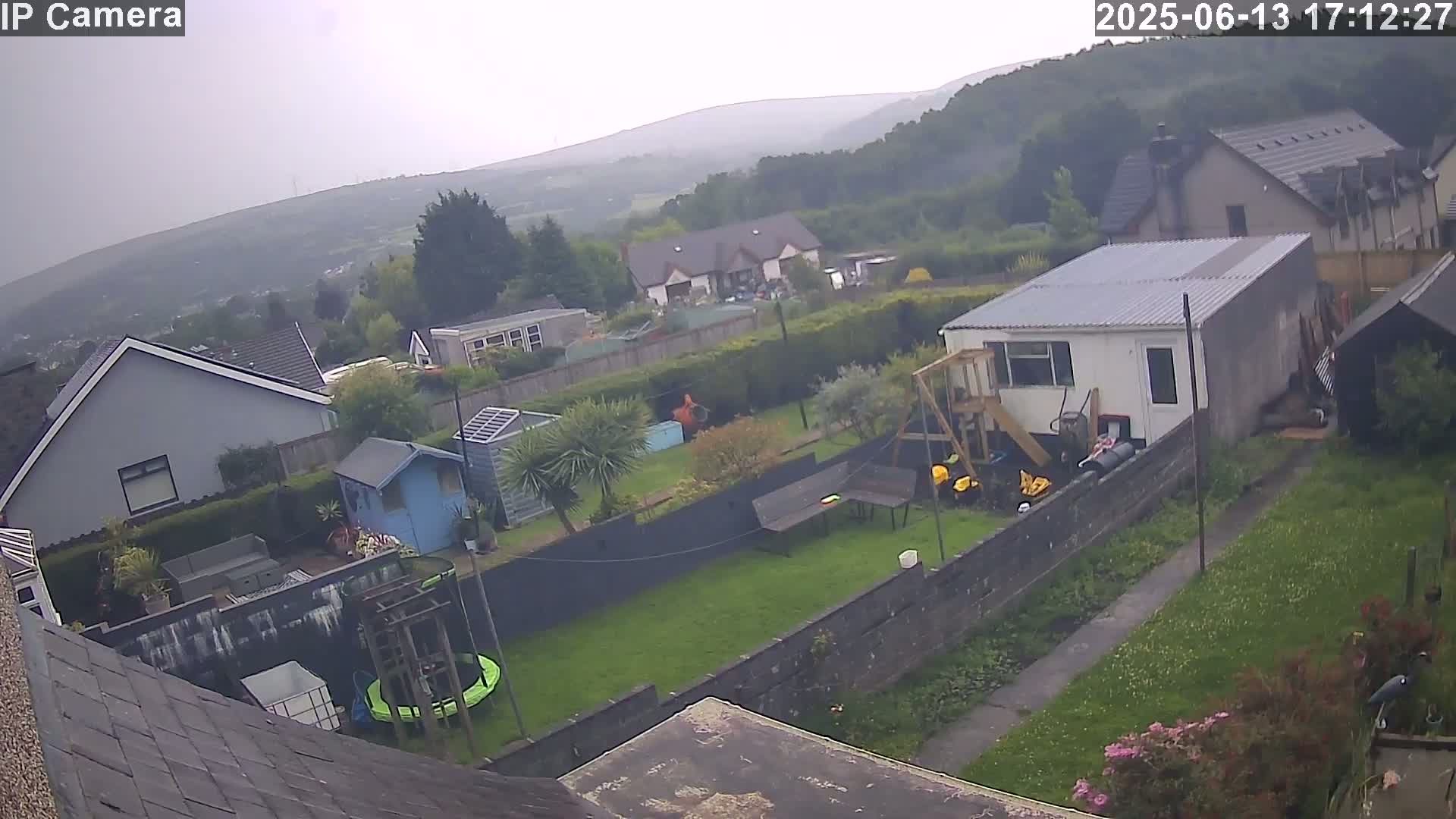 An overcast view shows a residential backyard with houses and hills in the distance, including a trampoline, playset, and garden shed.