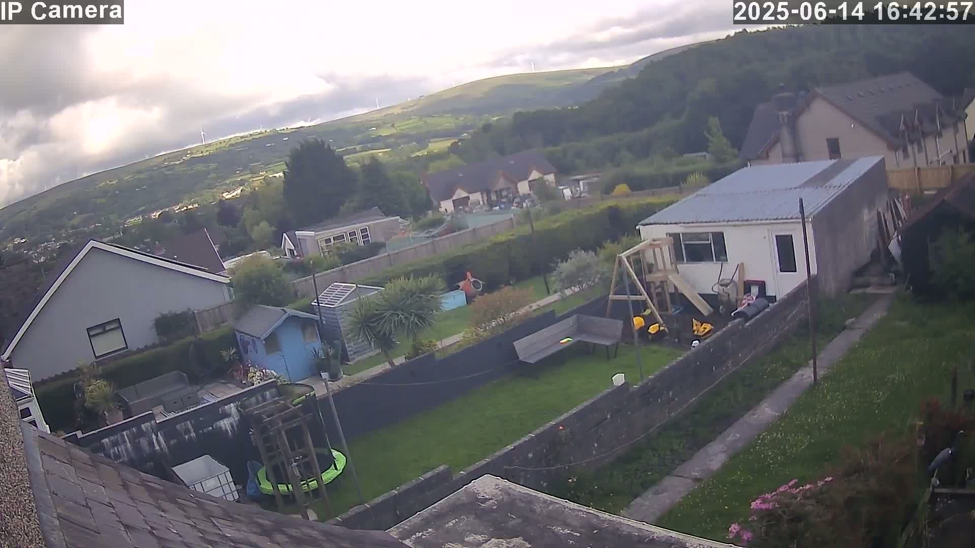 An overcast sky overlooks a residential backyard with a trampoline, playset, and several outbuildings, situated amongst other houses and green hills in the distance.
