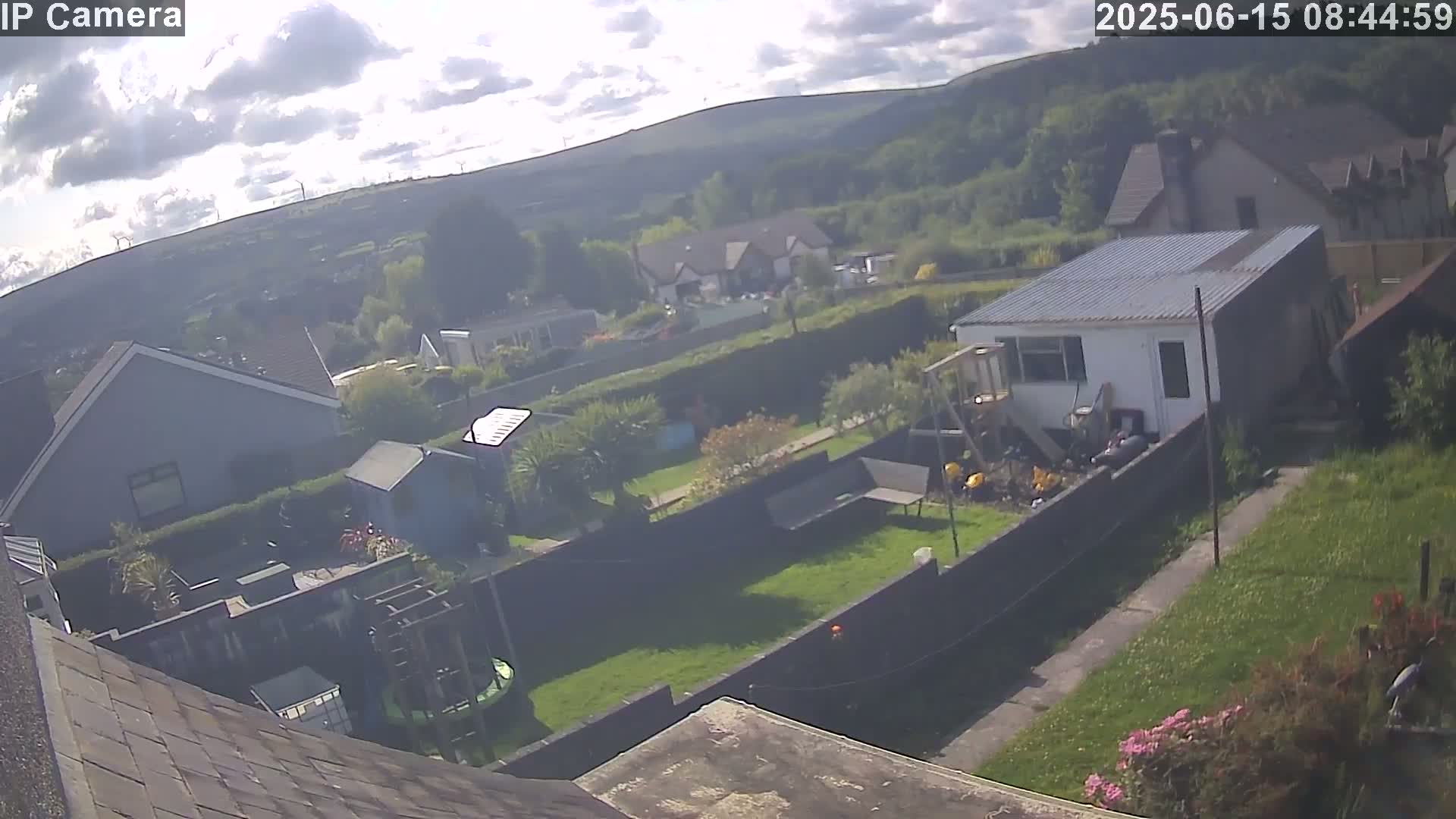 A high-angle view of a residential backyard on a partly sunny day, showing houses, gardens, and a small shed, with hills visible in the background.