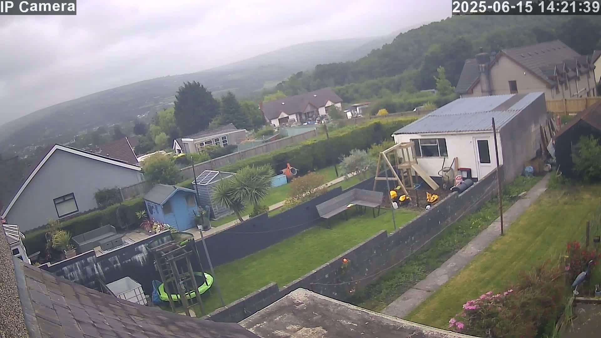 An overcast day reveals a backyard scene with houses, a shed, a playset, a trampoline, and a walkway leading to a grassy area, all nestled amongst greenery with a hilly landscape in the background.
