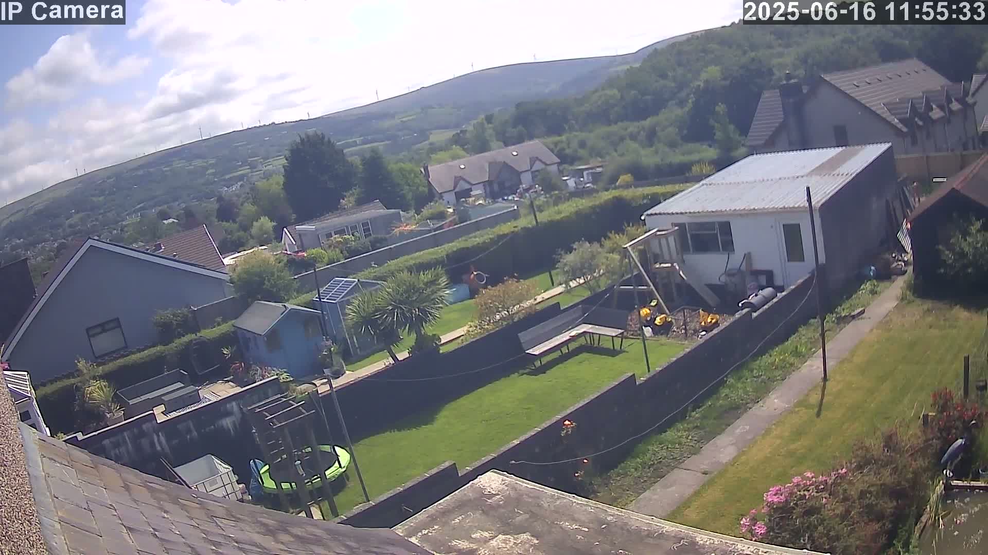 A partly cloudy day overlooks a backyard with a green lawn, various outbuildings, and a trampoline, all situated against a backdrop of houses and rolling hills dotted with wind turbines.