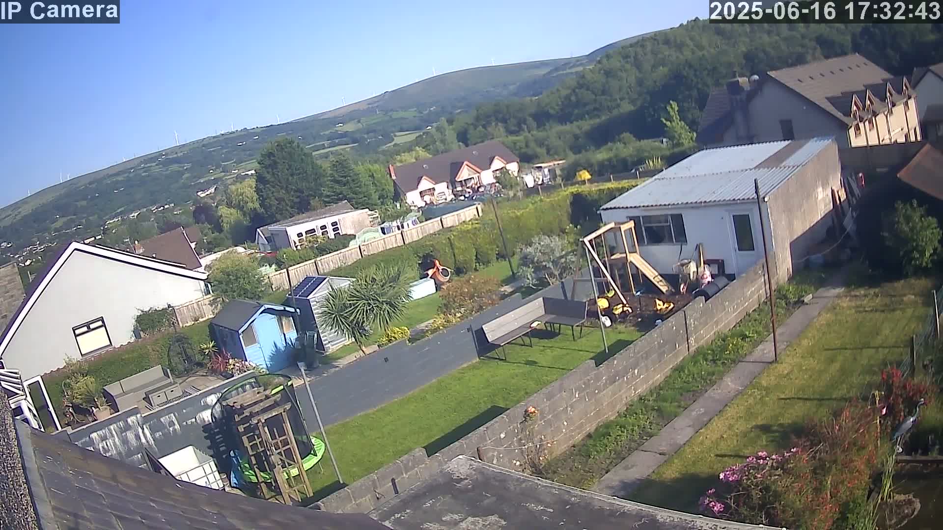 A backyard scene on a sunny day shows houses, a grassy area with a playground, a trampoline, and a garden, all overlooking a valley and distant hills.