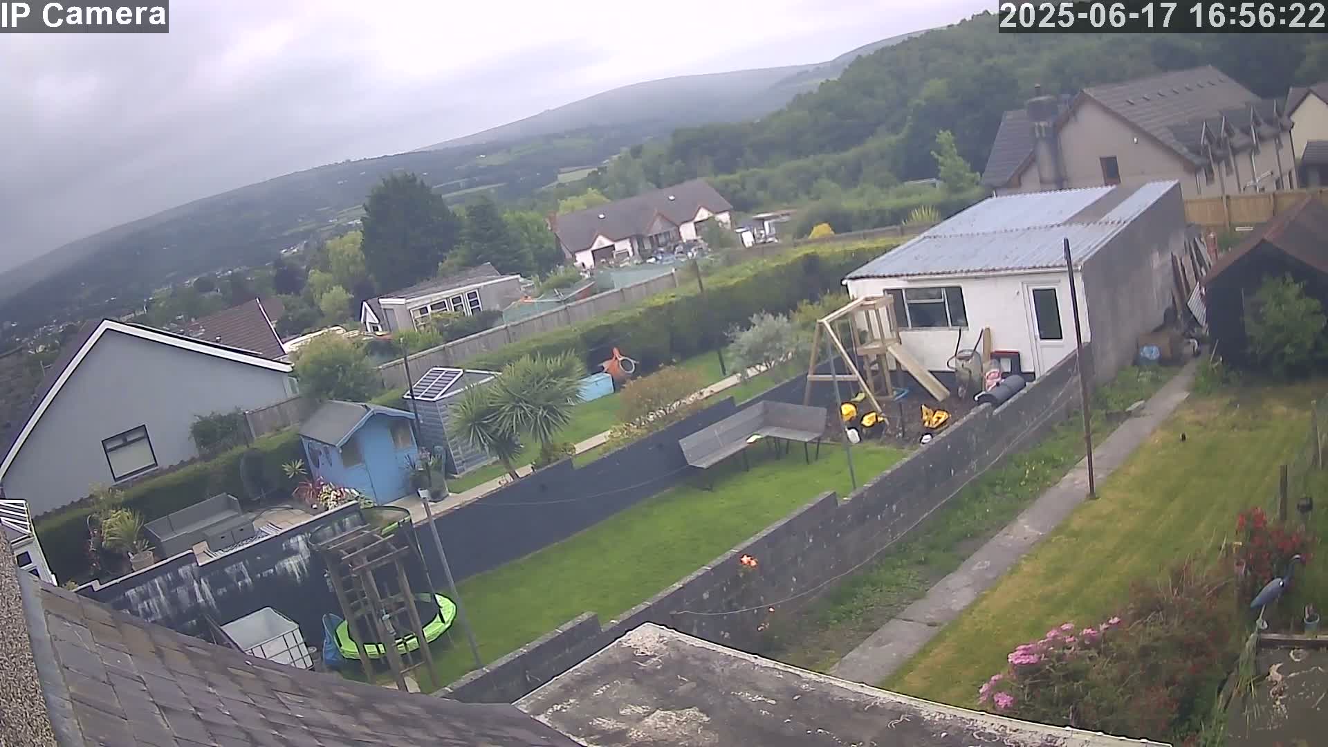 An overcast day reveals a backyard with houses and greenery in the background, a garden with a trampoline and playset, and a small building.