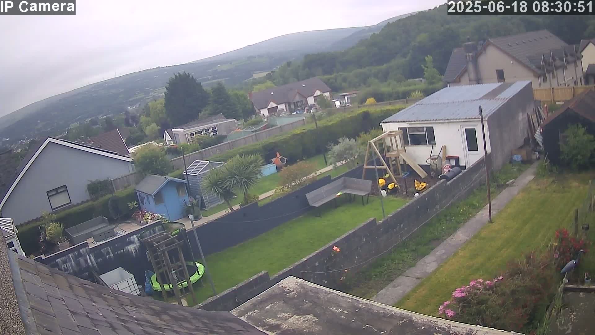 A backyard scene on an overcast day shows a green lawn, a small building, a trampoline, a playground set, and several houses nestled in a hilly, wooded area.