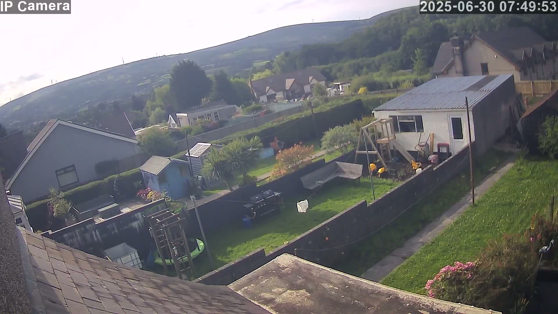 A high-angle view shows a backyard with houses and hills in the background under partly cloudy skies.